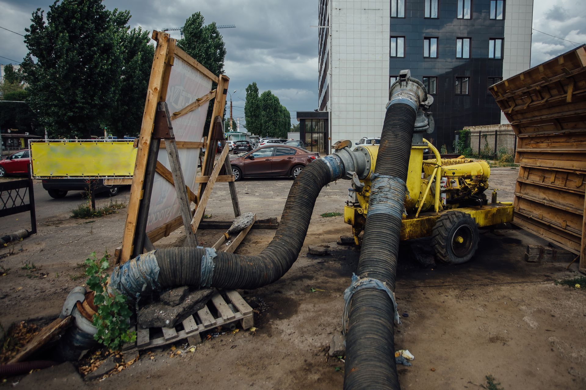 Construction site with large black ventilation ducts, yellow machinery, and stacked materials near buildings