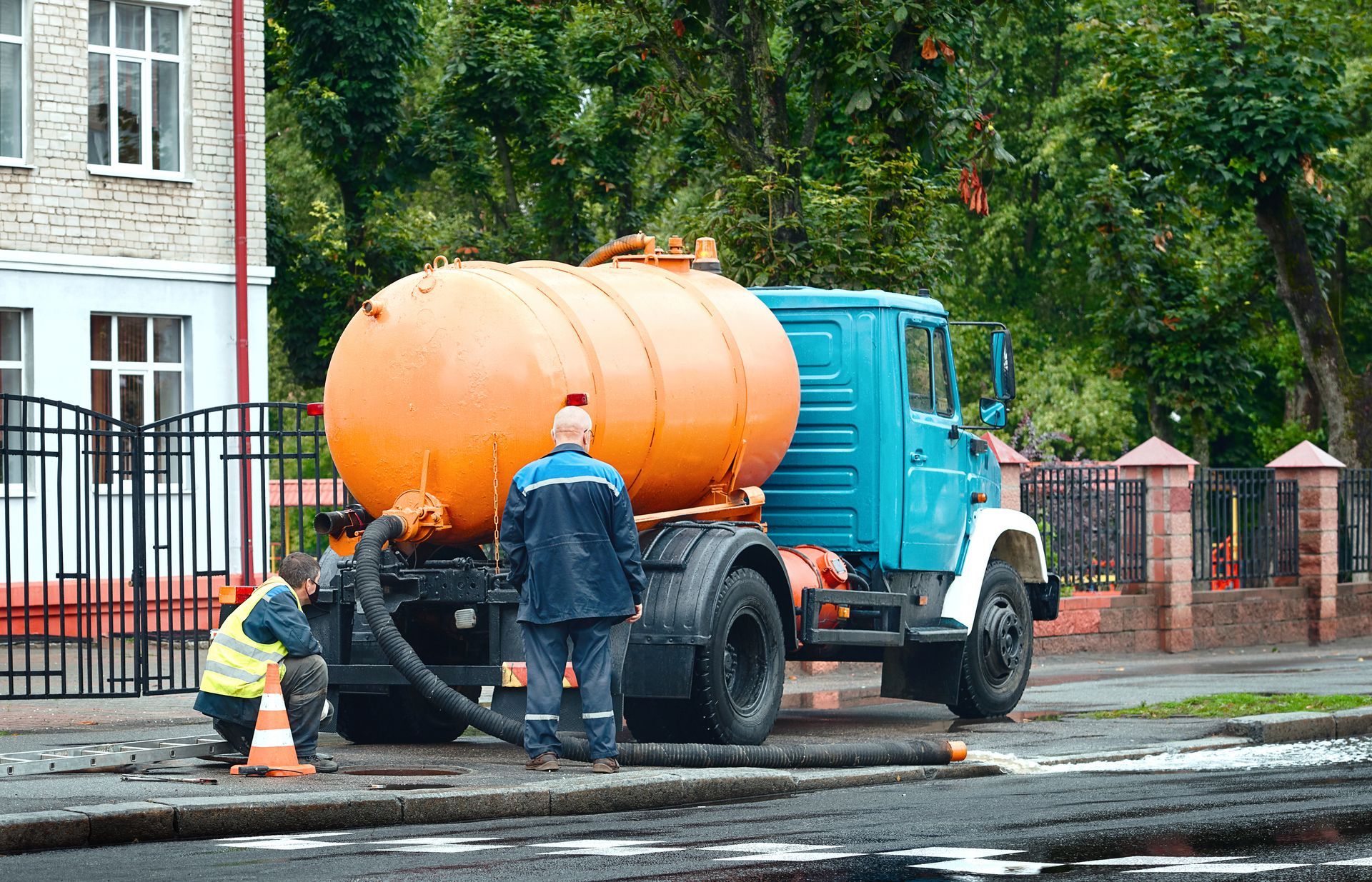 Orange tanker truck on a wet street with two workers inspecting it near a building