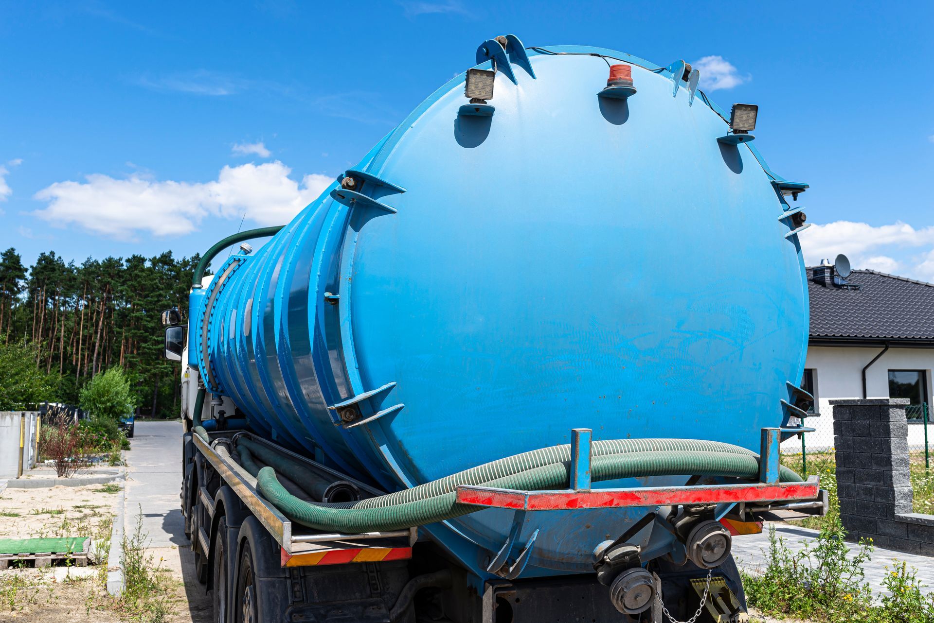 Blue tanker truck with a large cylindrical tank parked outdoors near houses and trees
