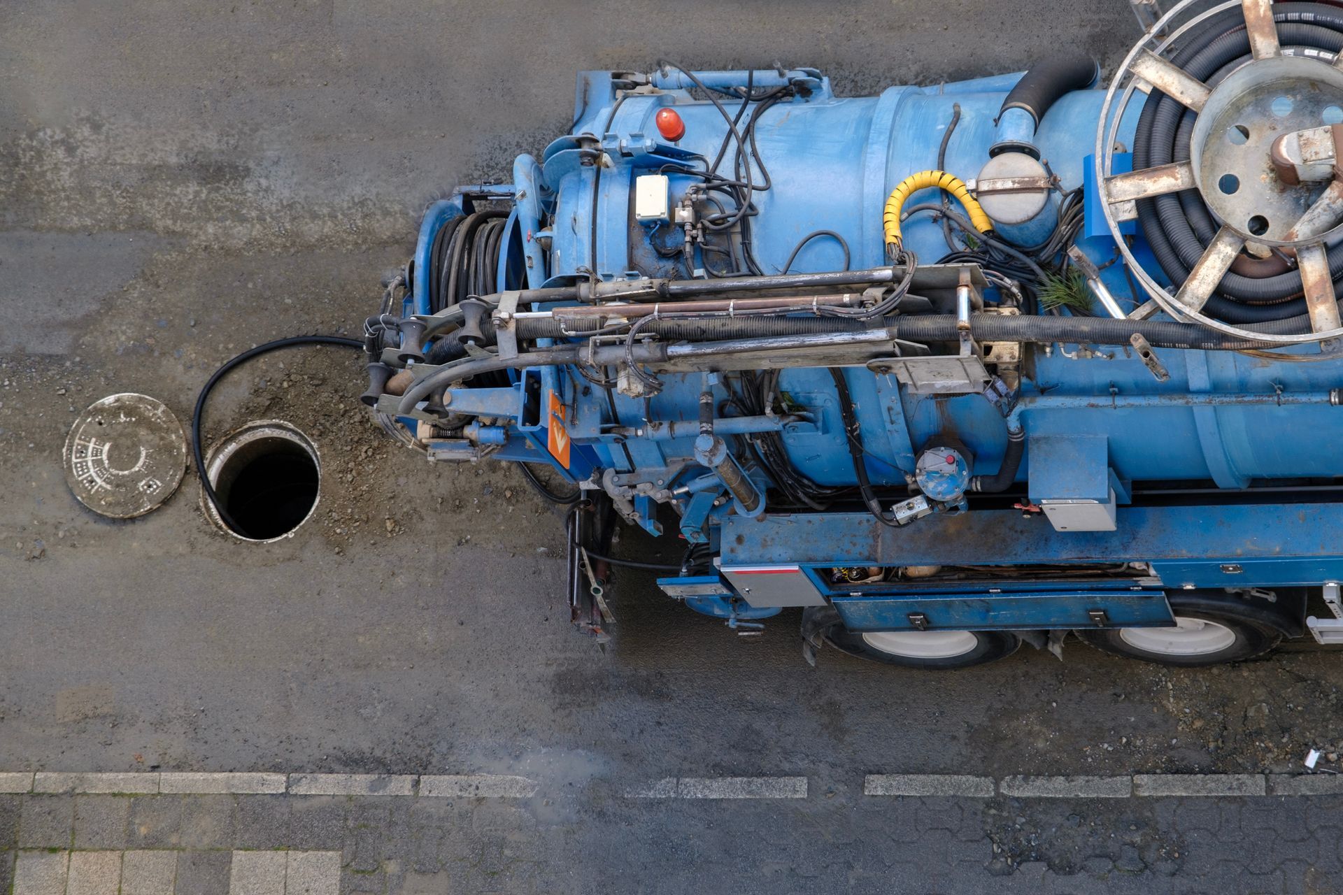 Blue industrial sewer-cleaning truck beside an open manhole on a wet street overhead view