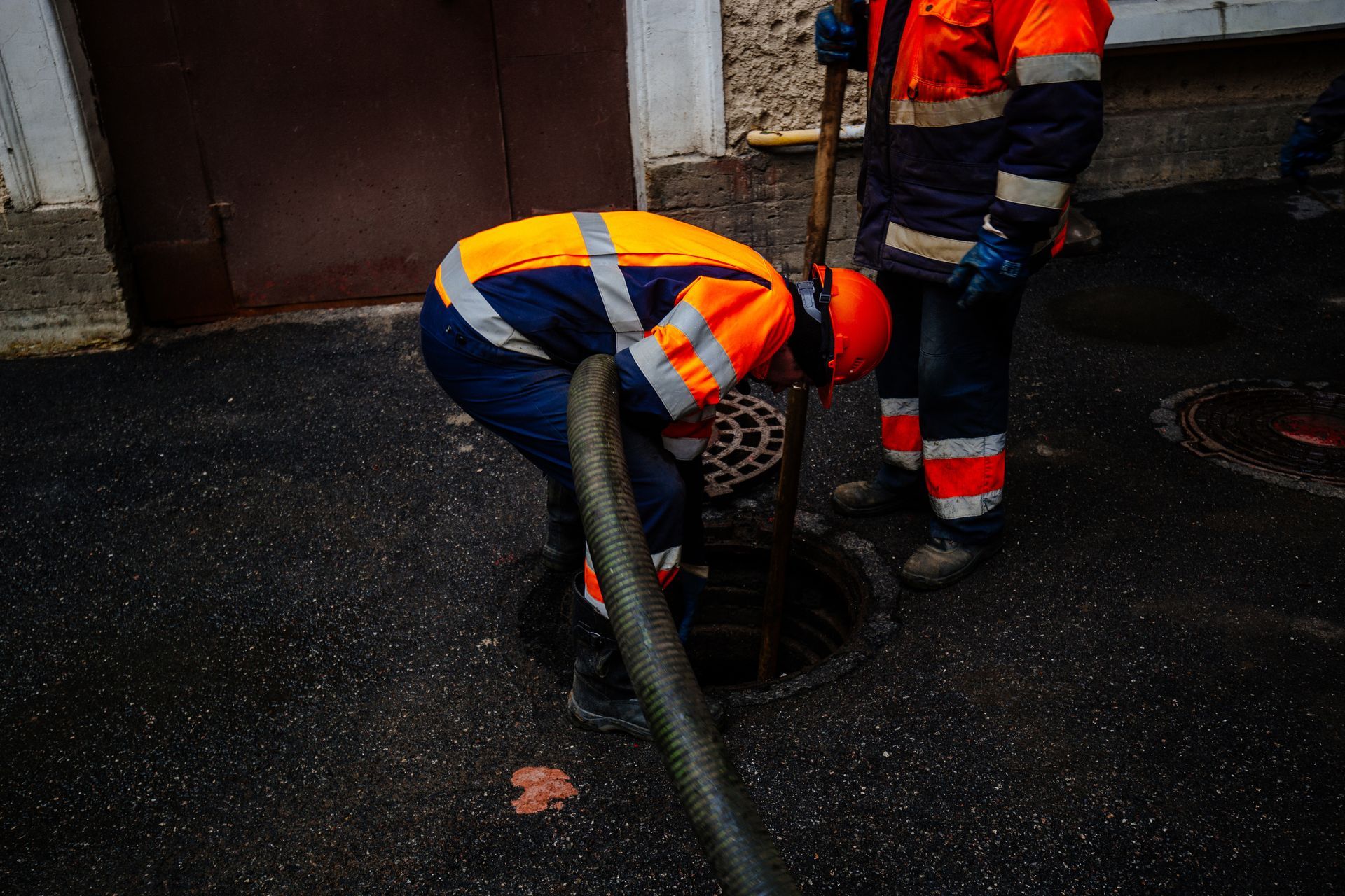 Two workers in orange safety gear inspect a hose on wet pavement by a building wall.