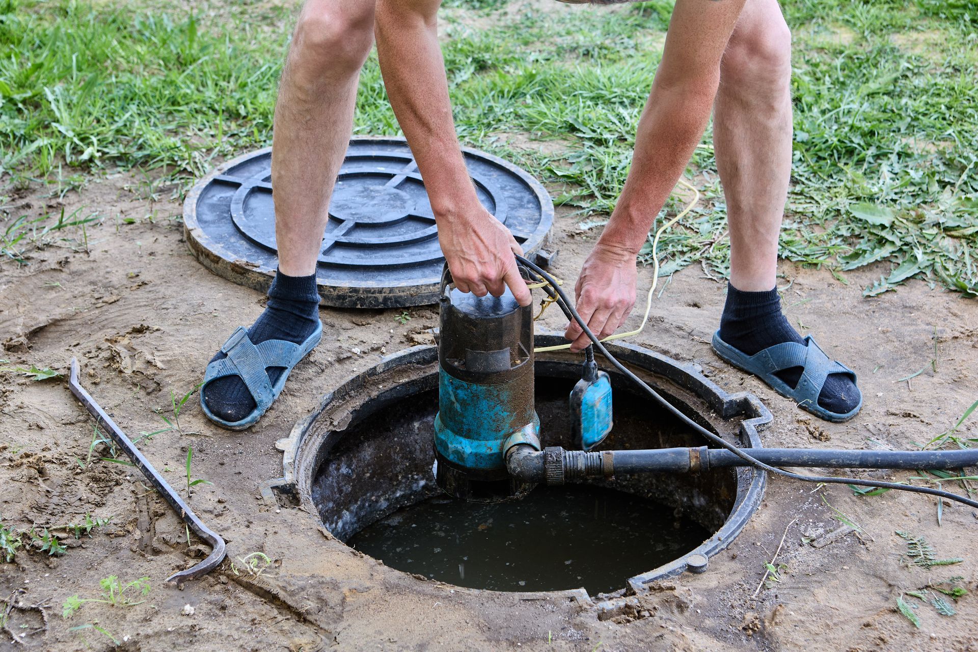 Two people working on an open manhole, lowering a tool into the circular sewer opening.