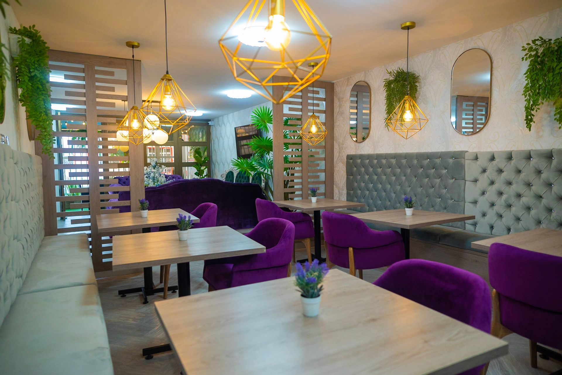 Un interior de cafetería con mesas de madera, sillas de terciopelo morado, modernas luces colgantes geométricas doradas y vegetación colgante.