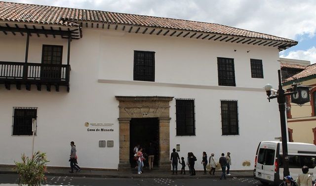 Un edificio histórico, blanco, de dos pisos, con balcones de madera oscura, una entrada con marco de piedra y gente caminando al frente.
