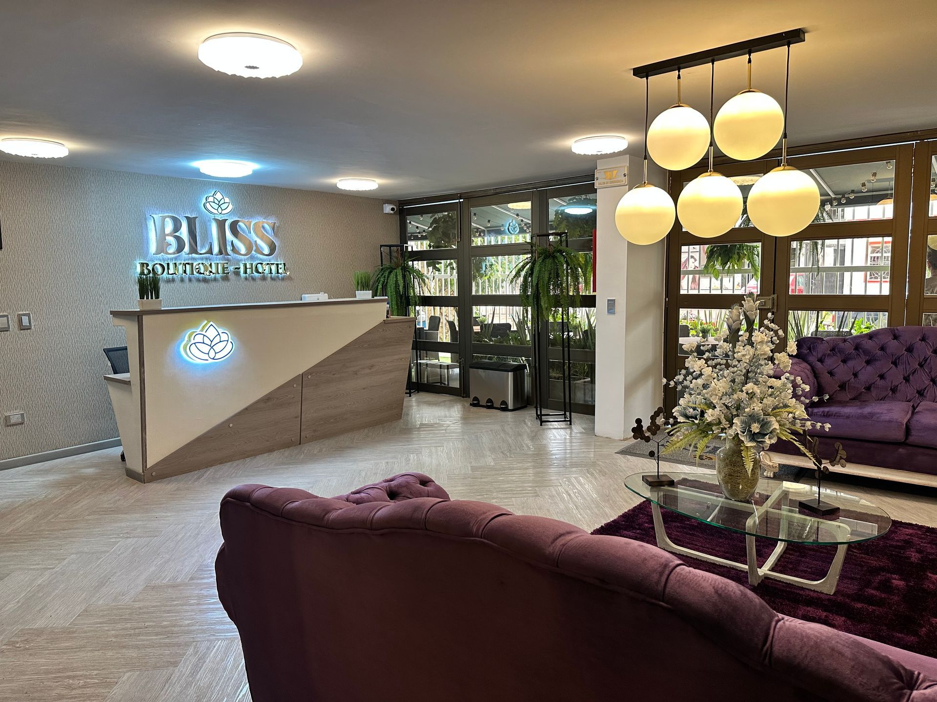 Vestíbulo de hotel moderno con mostrador de recepción, sofás con mechones morados, mesa de centro de cristal y luces colgantes con forma de globo.