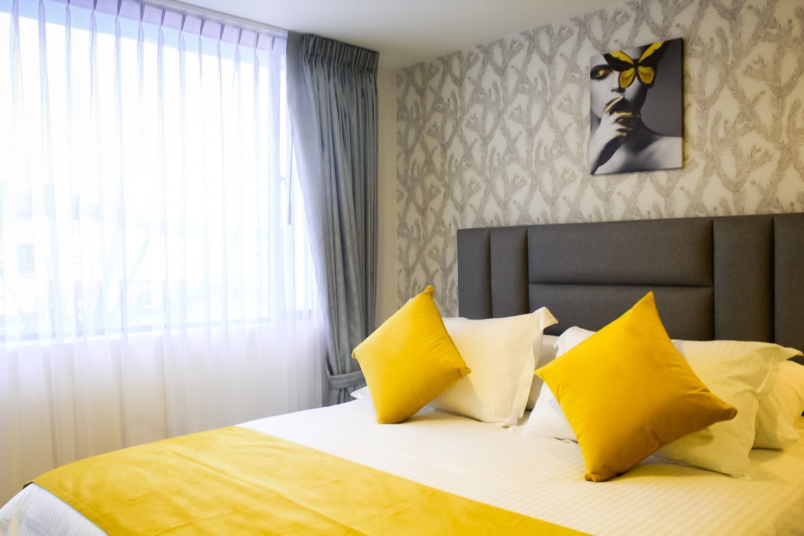 Un dormitorio de hotel moderno con cabecera capitoné gris, ropa de cama blanca con detalles amarillos y arte de pared con mariposas.