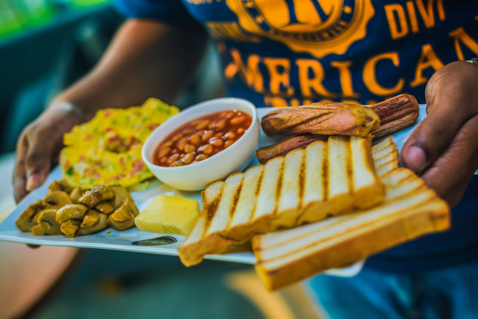 Una persona sostiene un plato de desayuno con una tortilla, frijoles en un tazón pequeño, salchichas a la parrilla, tostadas y champiñones salteados.