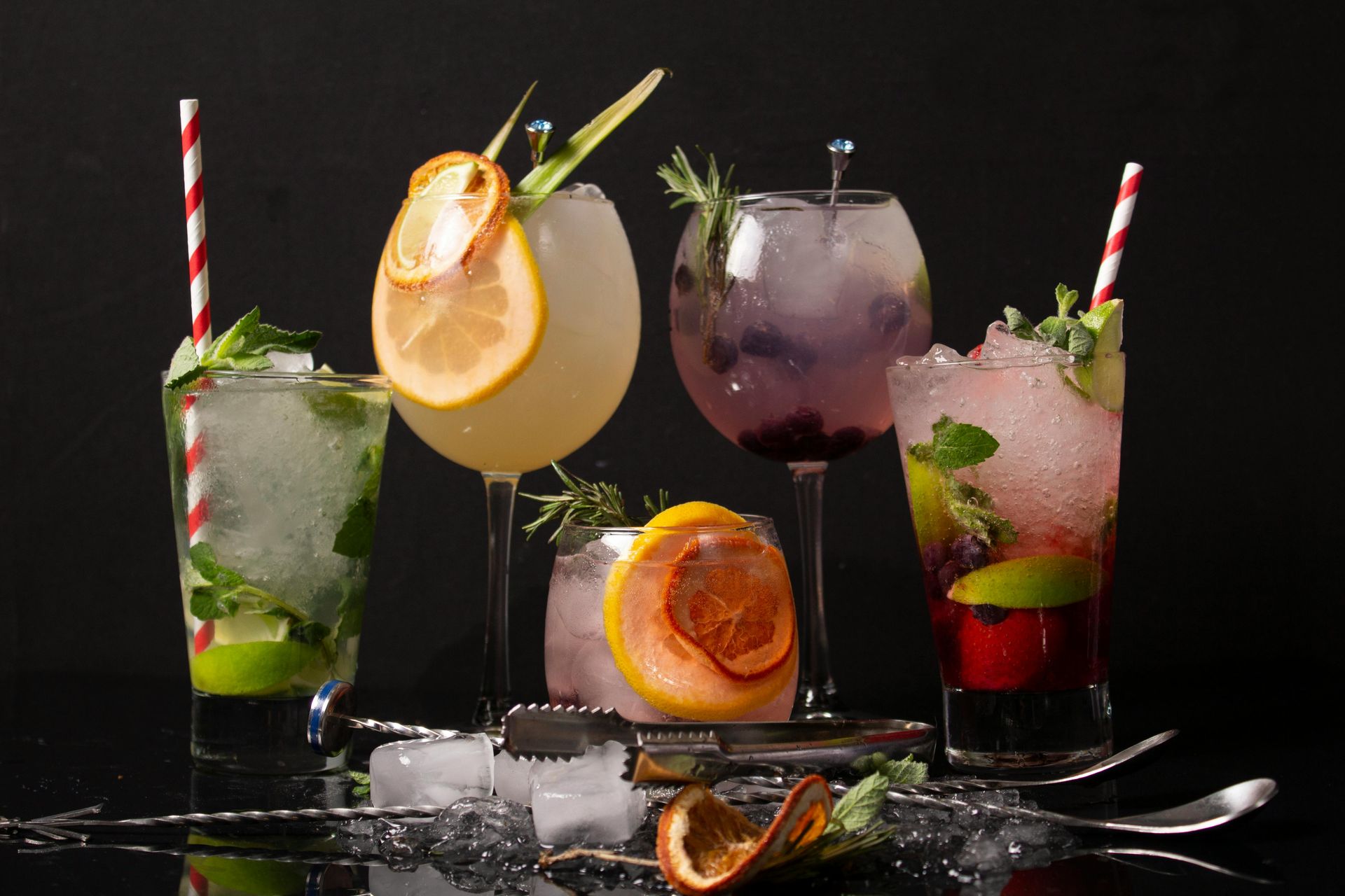 Cinco cócteles coloridos adornados con frutas y hierbas, dispuestos sobre una superficie oscura y reflectante con hielo y herramientas de barra.