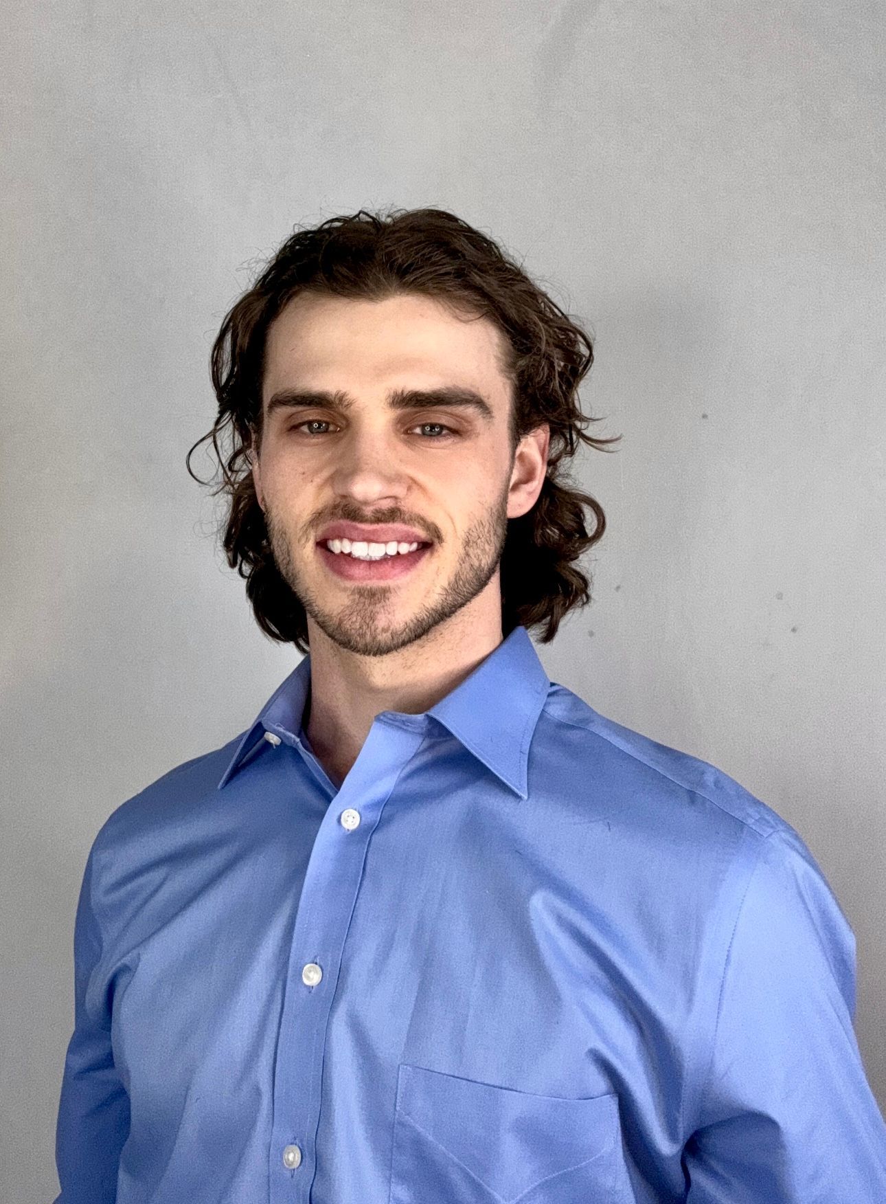 A man in a blue shirt is standing with his arms crossed and smiling.