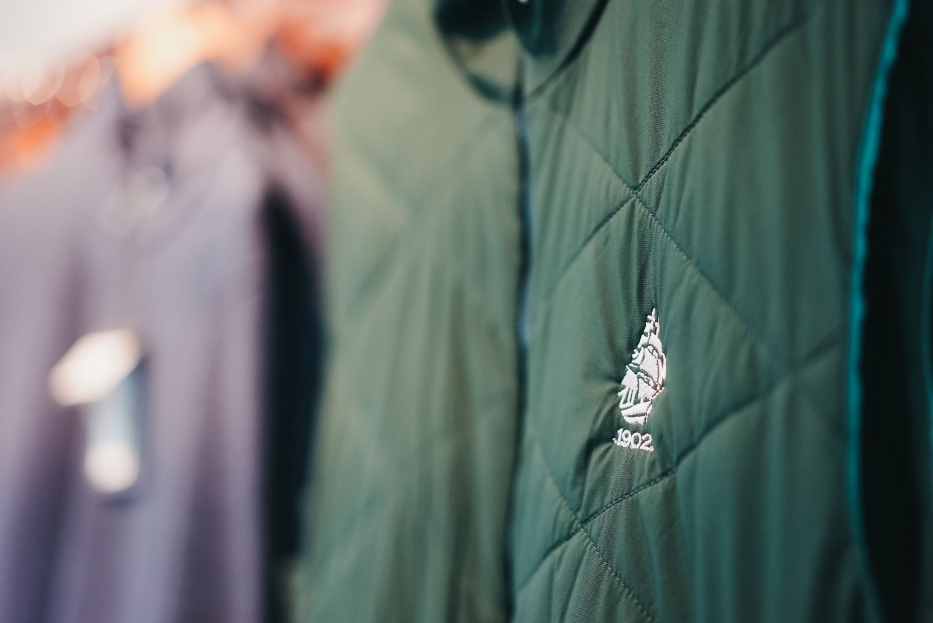 Green quilted jacket with a logo, hanging on a rack with other clothing.