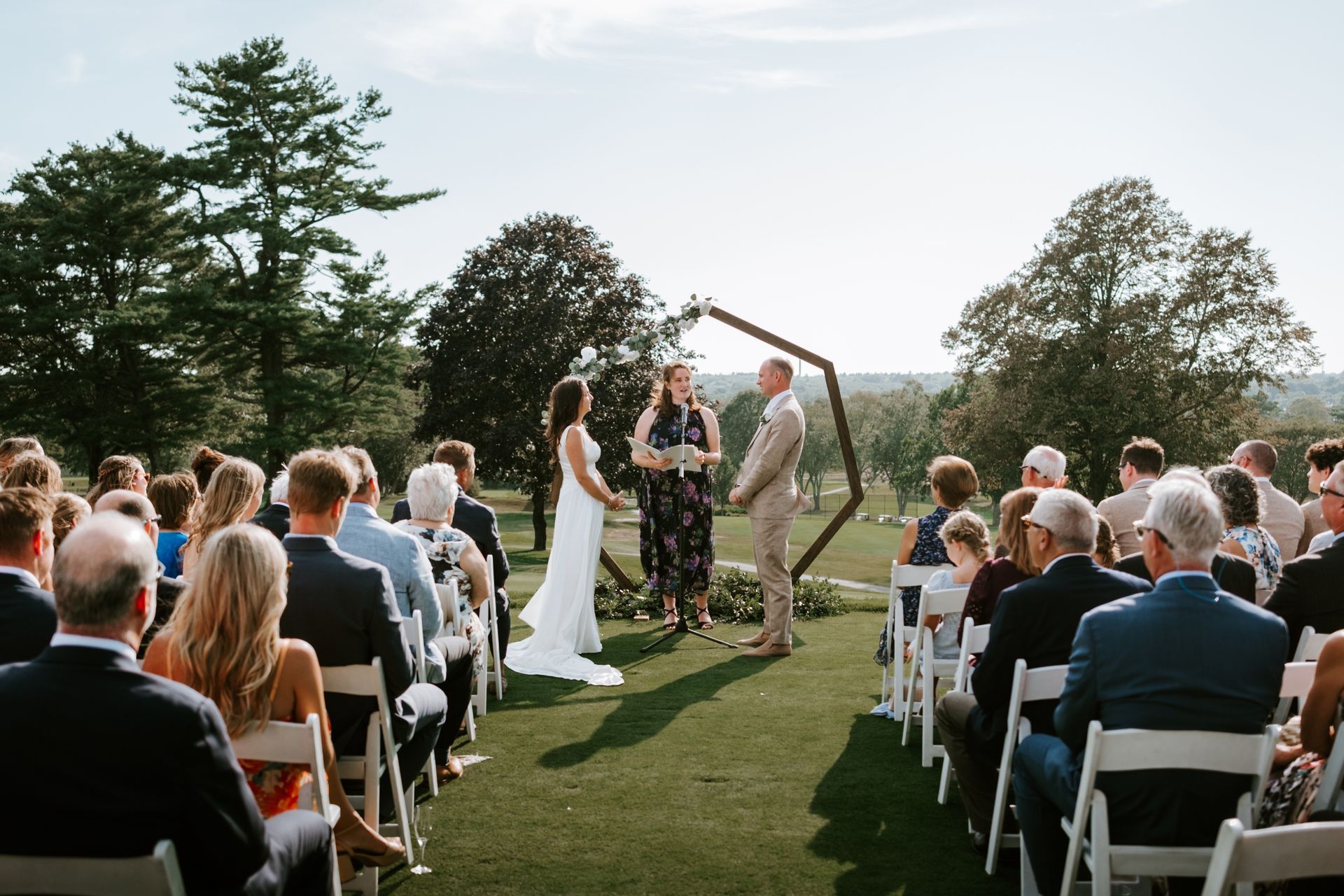 Wedding ceremony outdoors with couple, officiant under geometric arch, guests seated in rows on grass.