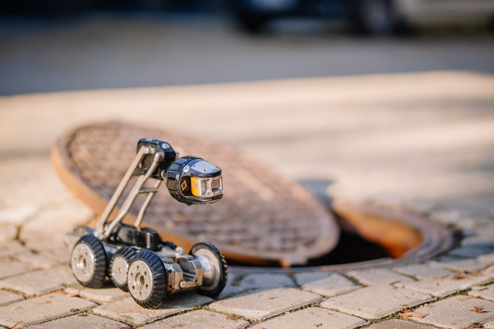 Small robot on a paved surface beside a manhole cover, with a blurred background.