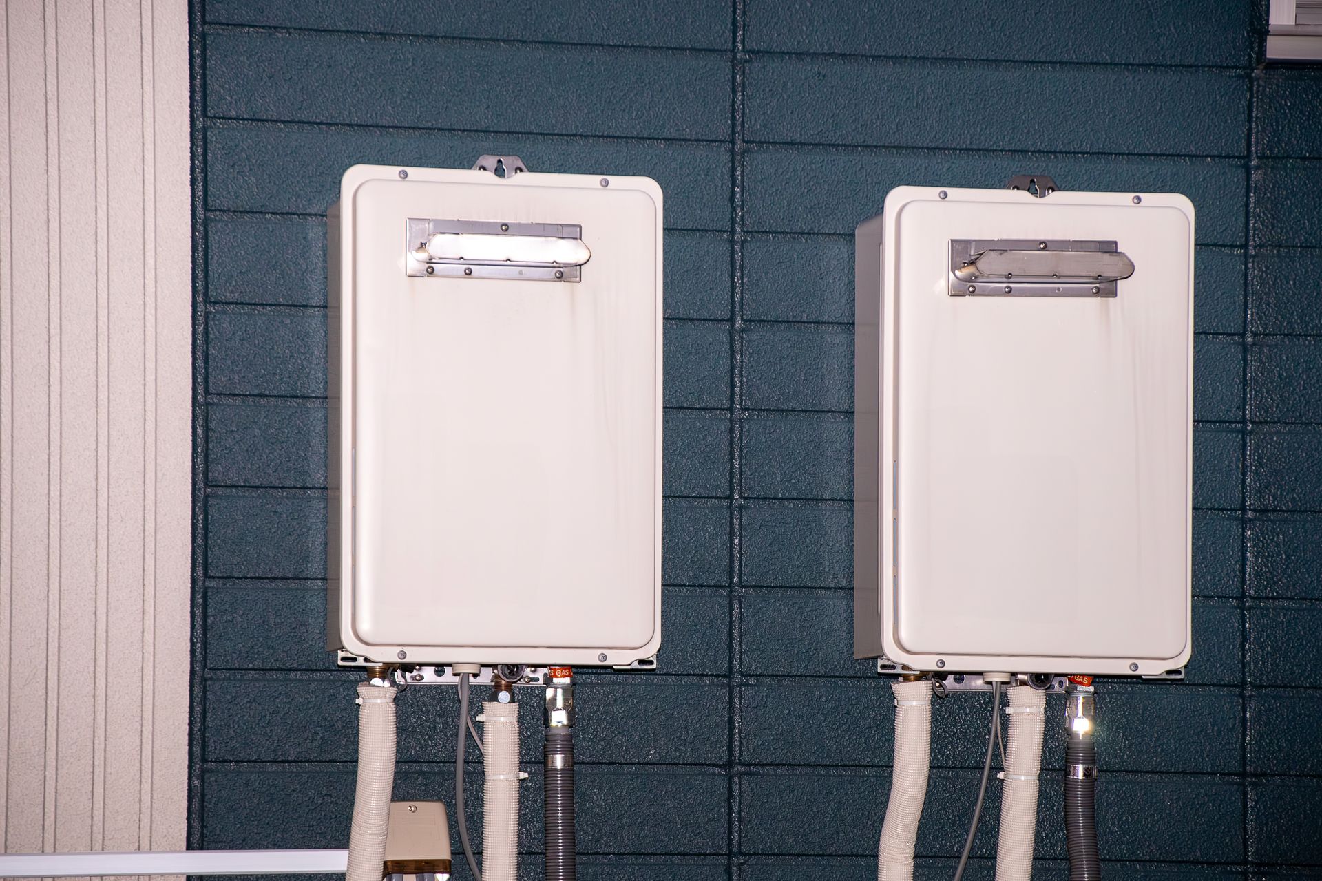 Two white utility boxes mounted on a dark brick wall with pipes and meters below