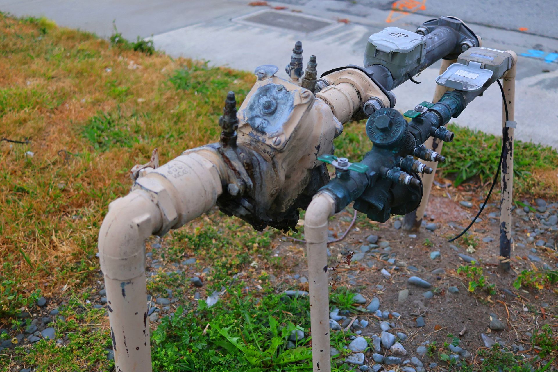 Rusty industrial pipes and valves with insulation, running through a grassy roadside area