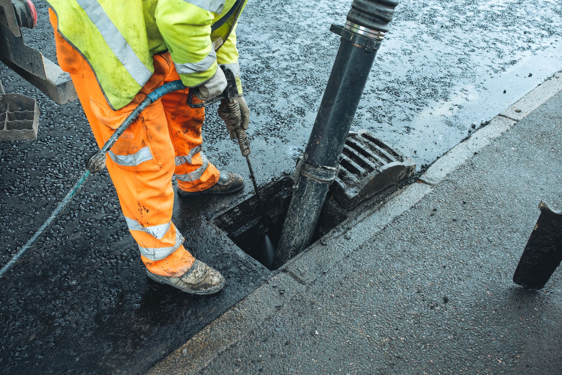 Roadworker in orange safety gear using a jackhammer to repair asphalt at a street excavation.