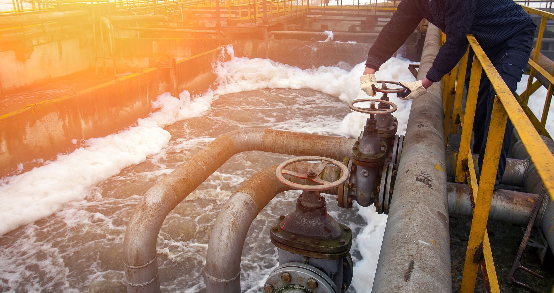 Worker operating a valve beside frothy water in an industrial pipe area, with yellow railings and sunrise light