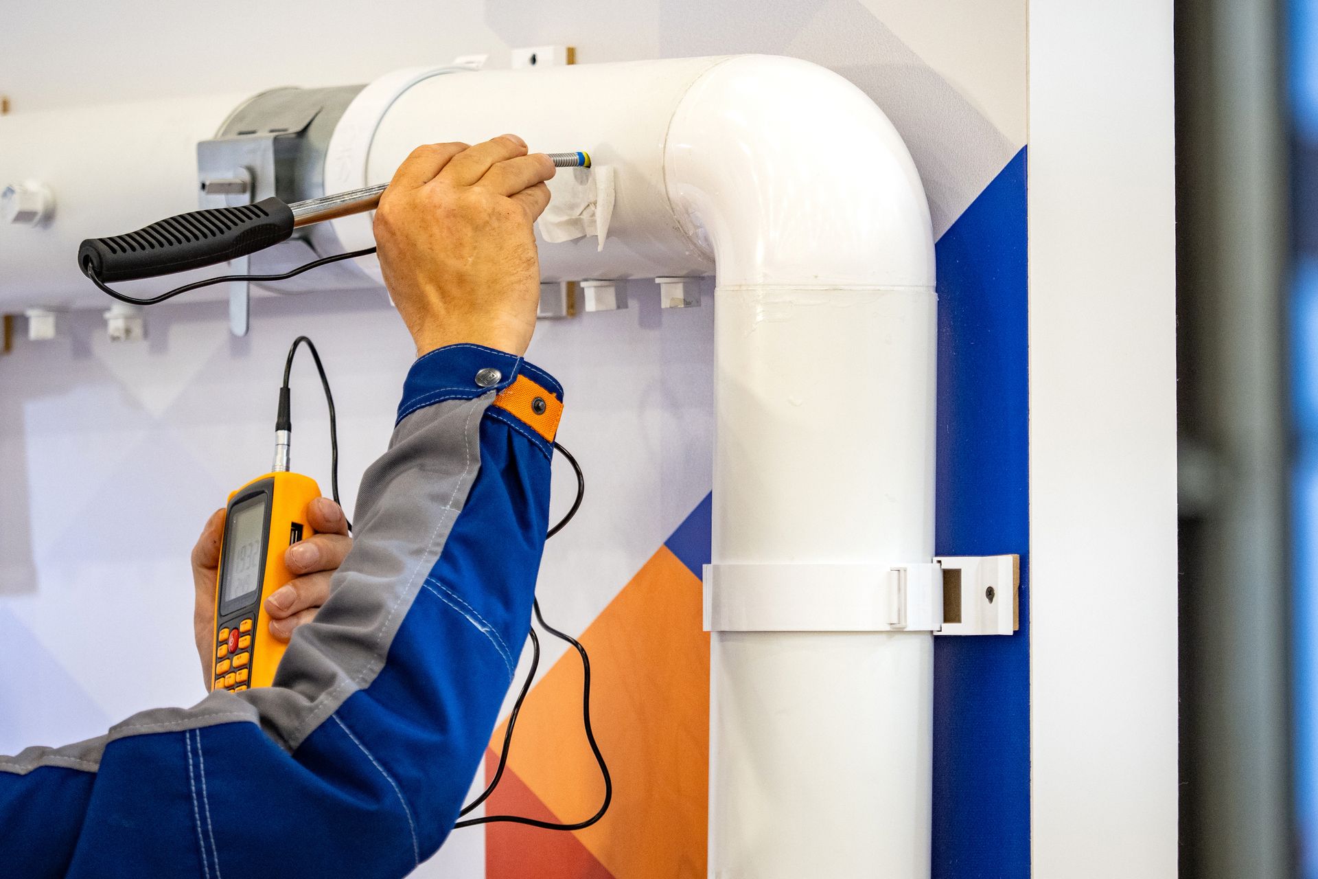 Technician tightening a pipe fitting with a drill near a white ventilation duct.