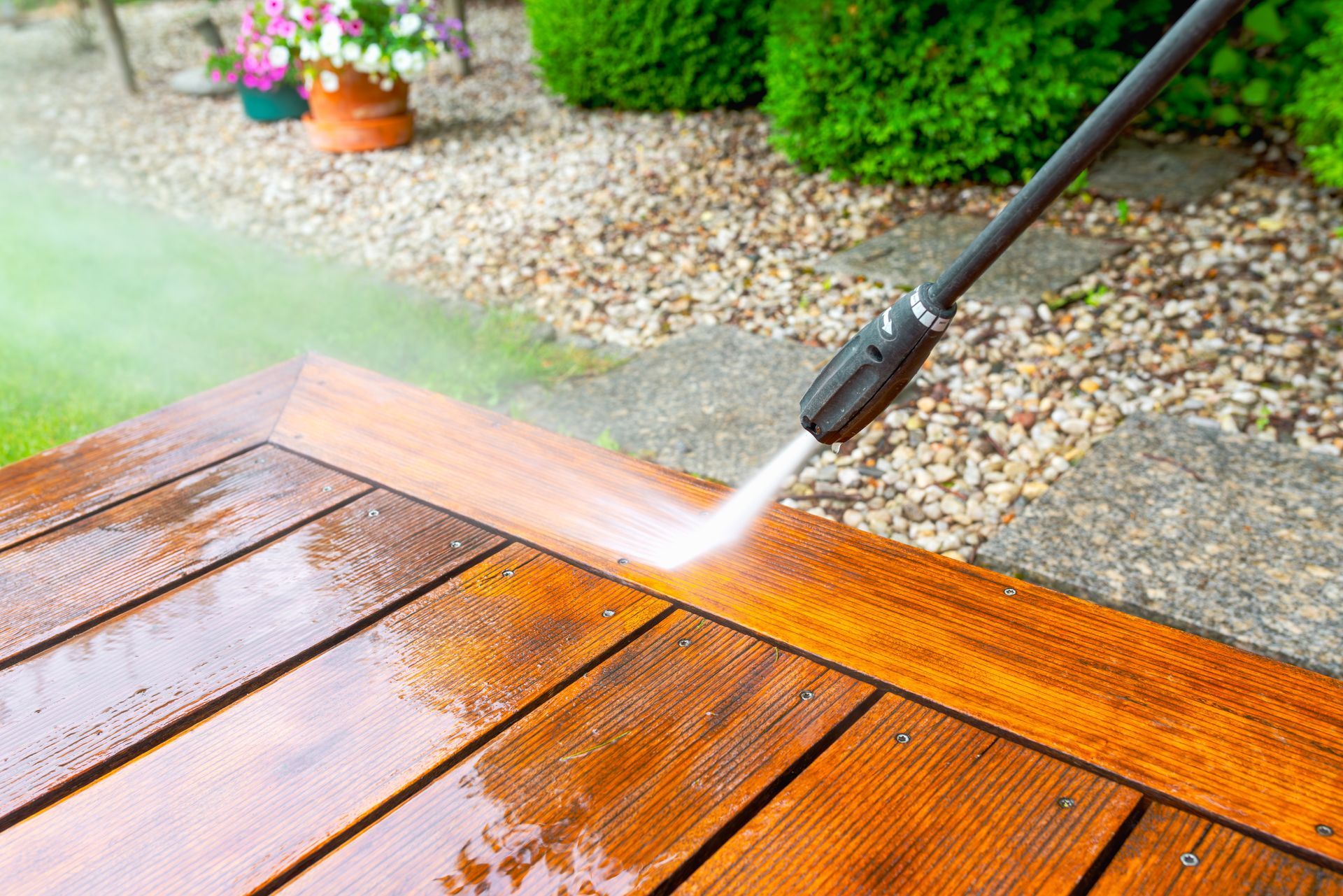 Pressure washer cleaning a wet wooden deck beside a gravel garden path