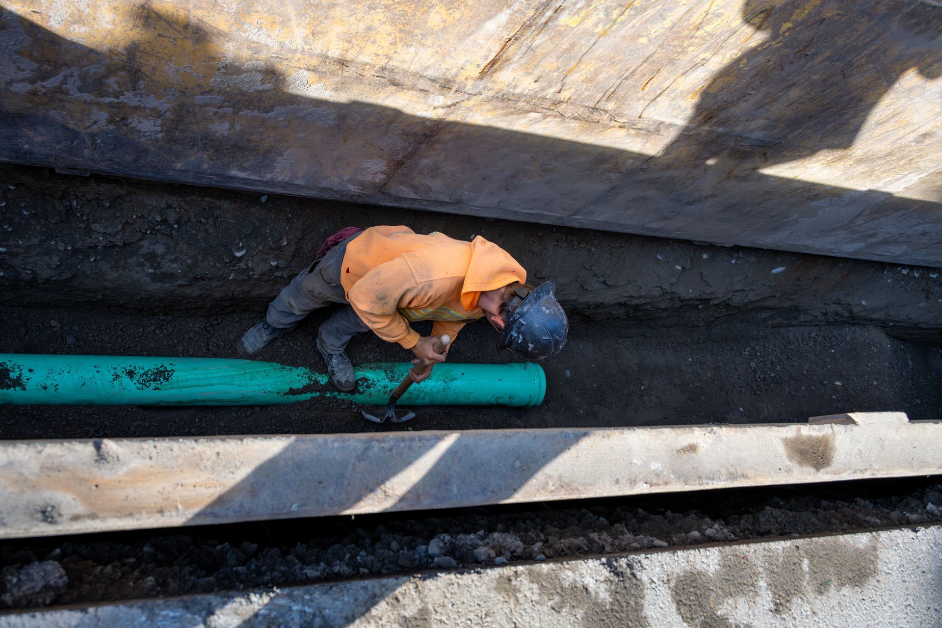 Worker in orange shirt repairing a green pipe inside a narrow concrete trench