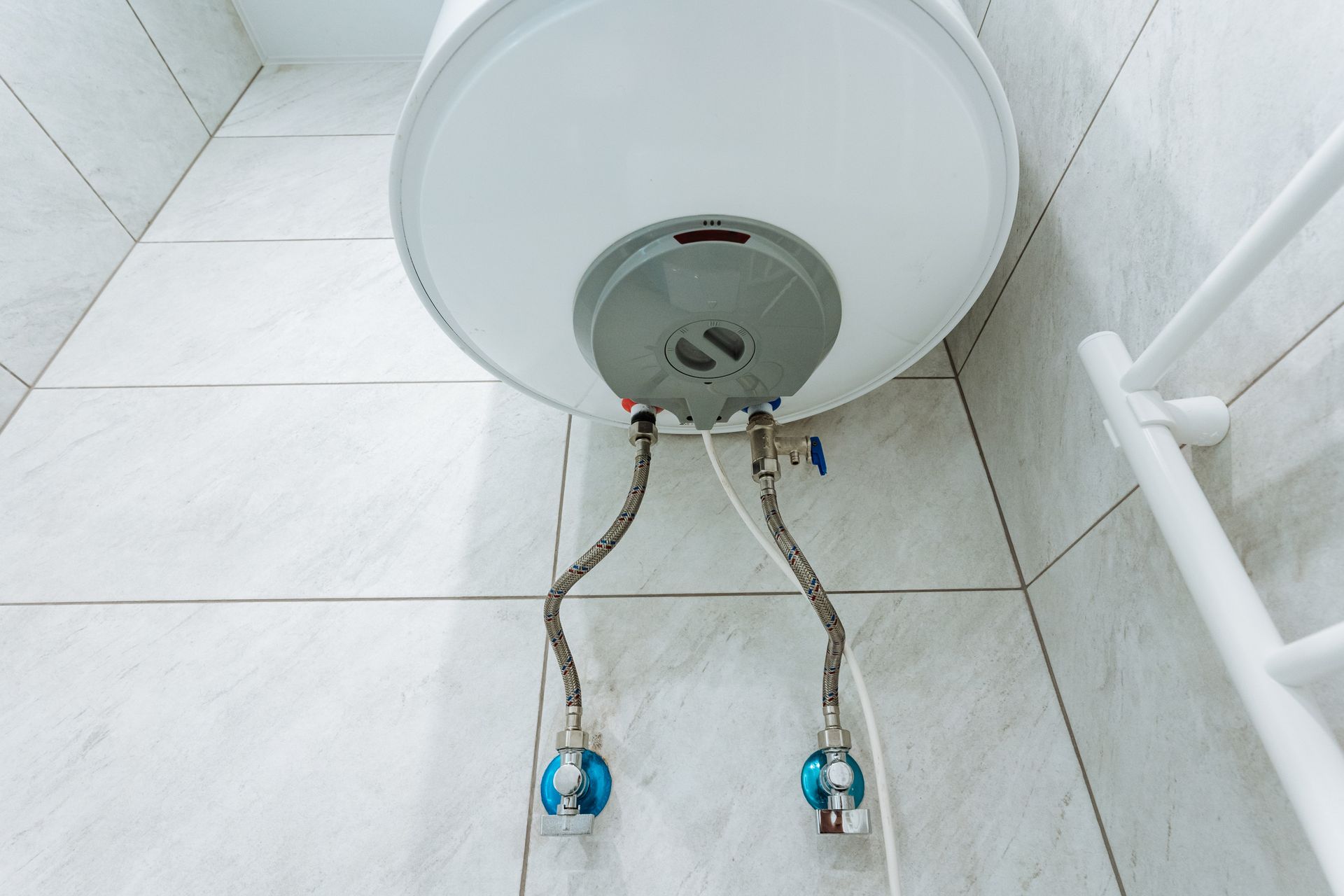Underside of a white water heater with exposed pipes and blue valves in a tiled bathroom