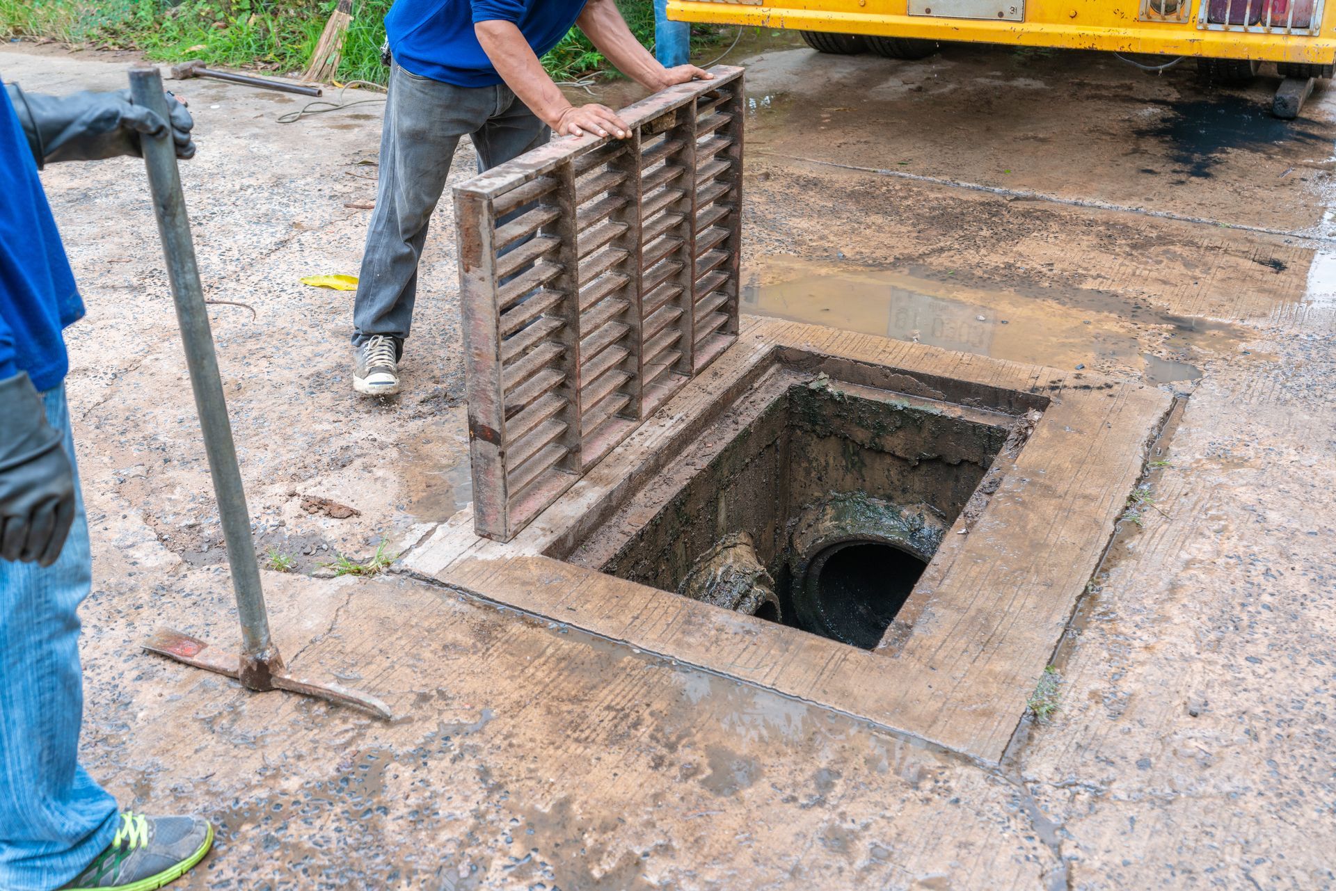 Worker lifting a metal grate over an open manhole on a muddy street lane