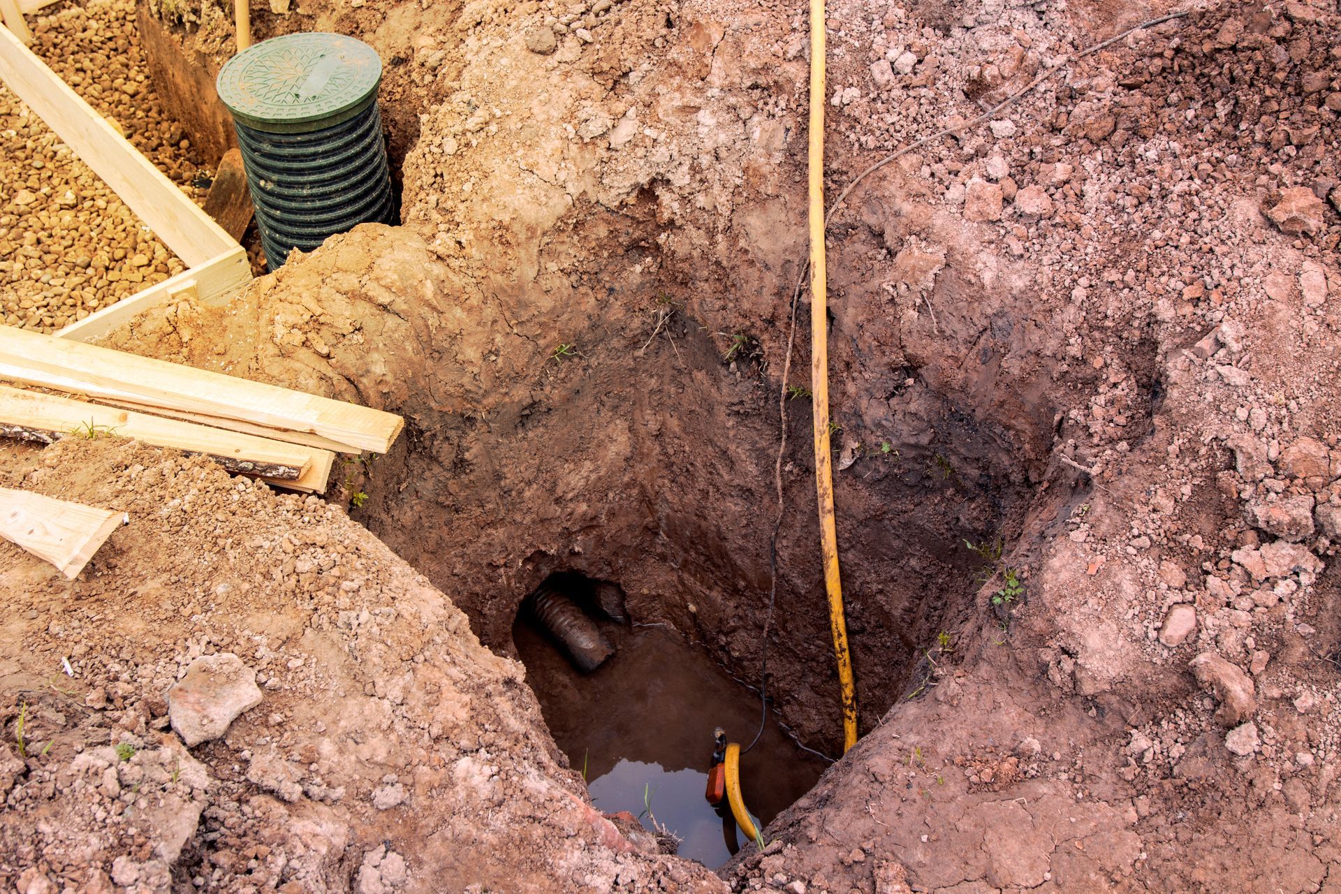 Construction trench with exposed dirt, wooden planks, and a yellow measuring line beside a pipe opening