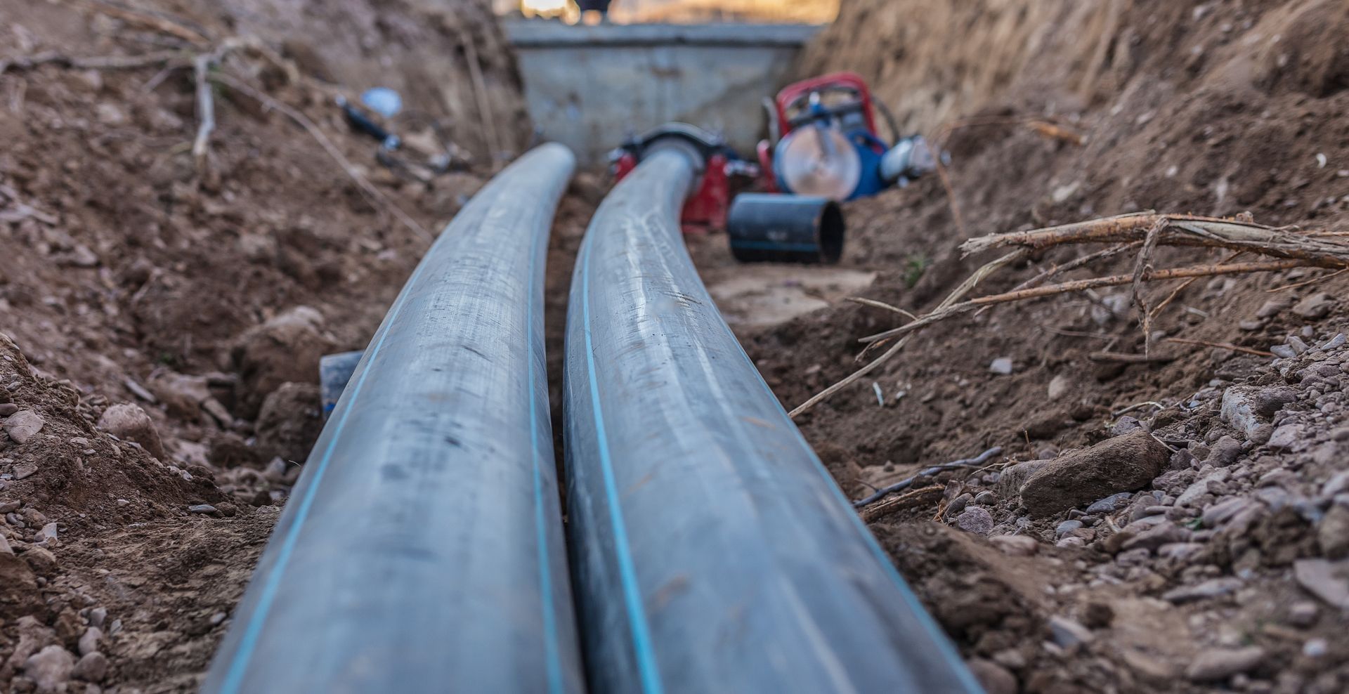 Two large black pipes laid in a dirt trench with construction equipment in the background