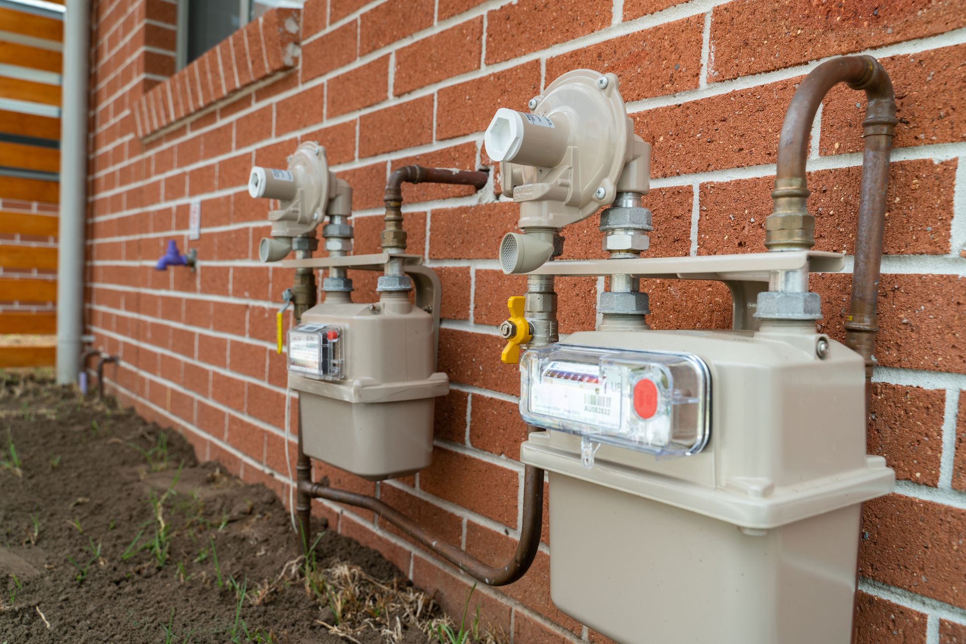 Two electric meters and utility boxes mounted on a red brick house exterior with pipes and cables.