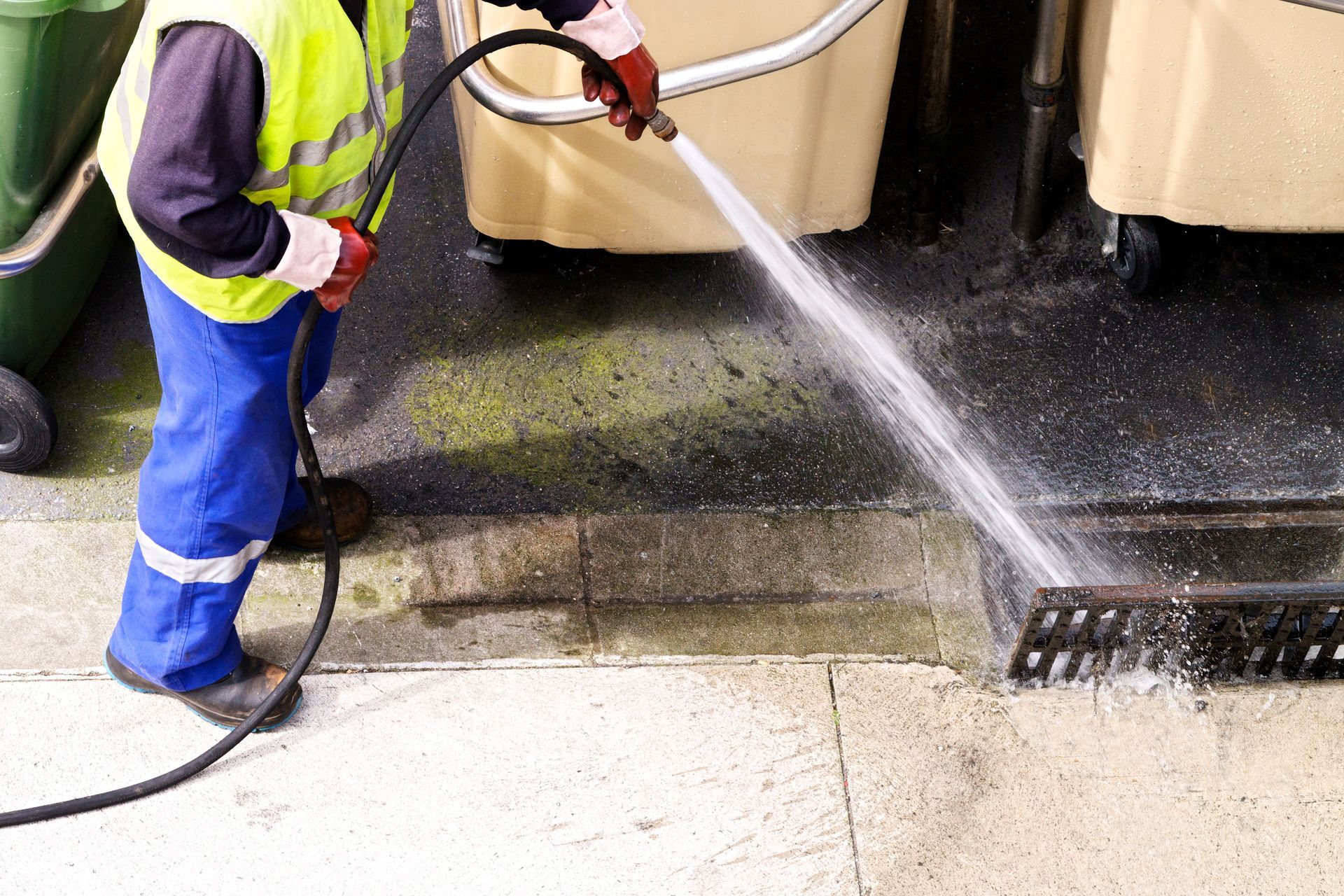 Worker pressure-washing a sidewalk beside large bins, spraying water onto the pavement