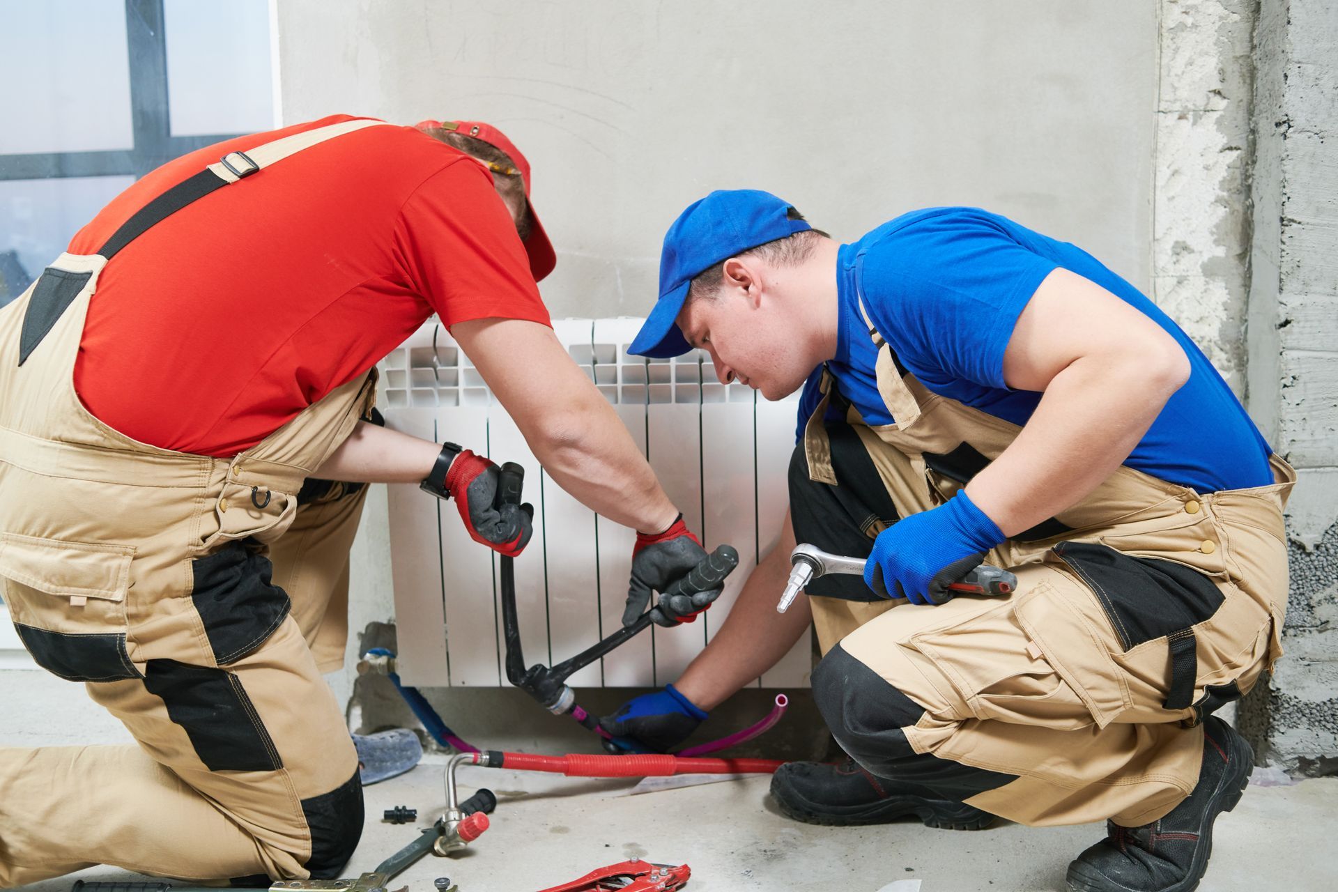 Two workers in red and blue shirts fixing a cable or pipe near a wall.
