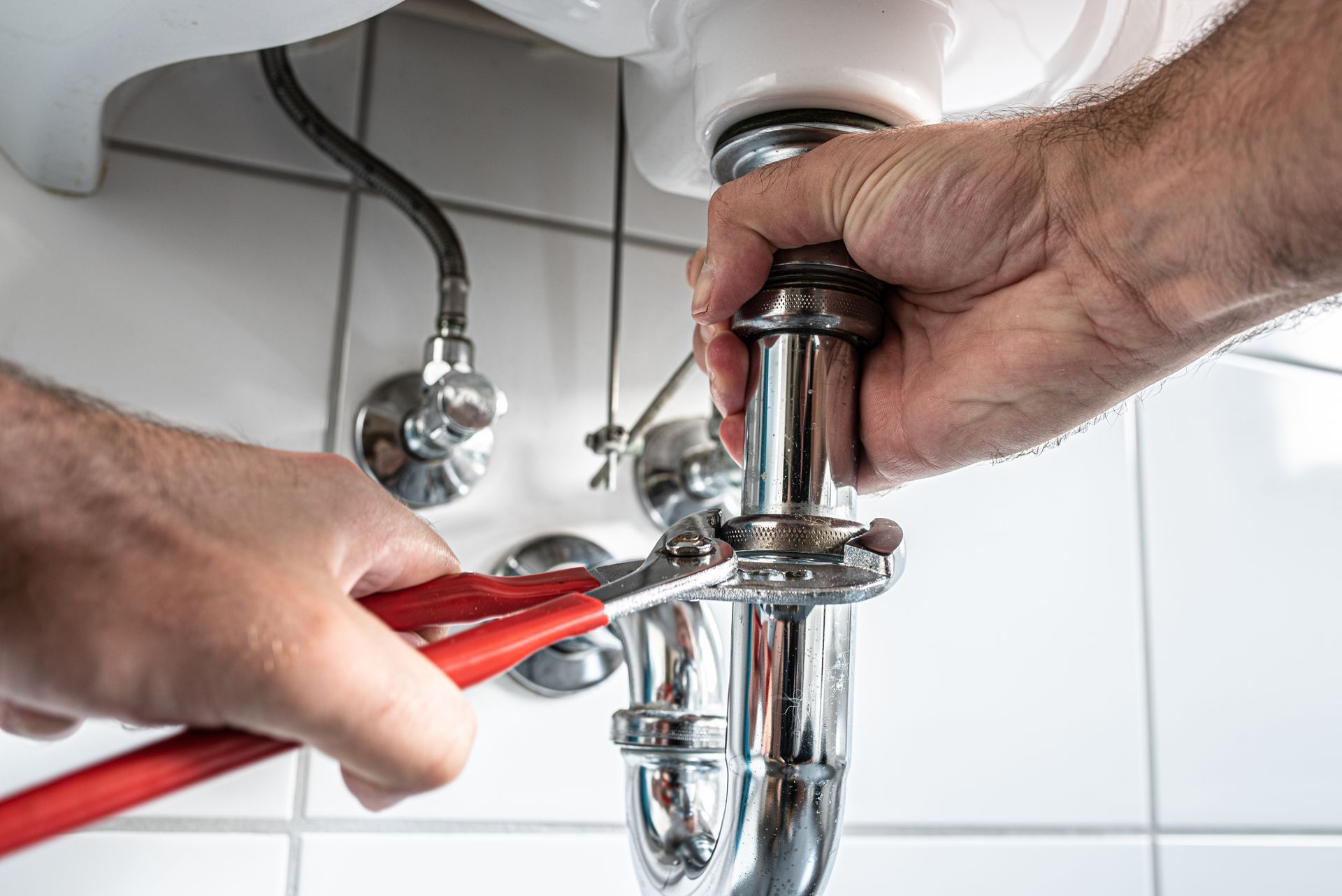 Hands using pliers to tighten a chrome sink drain pipe under a bathroom sink