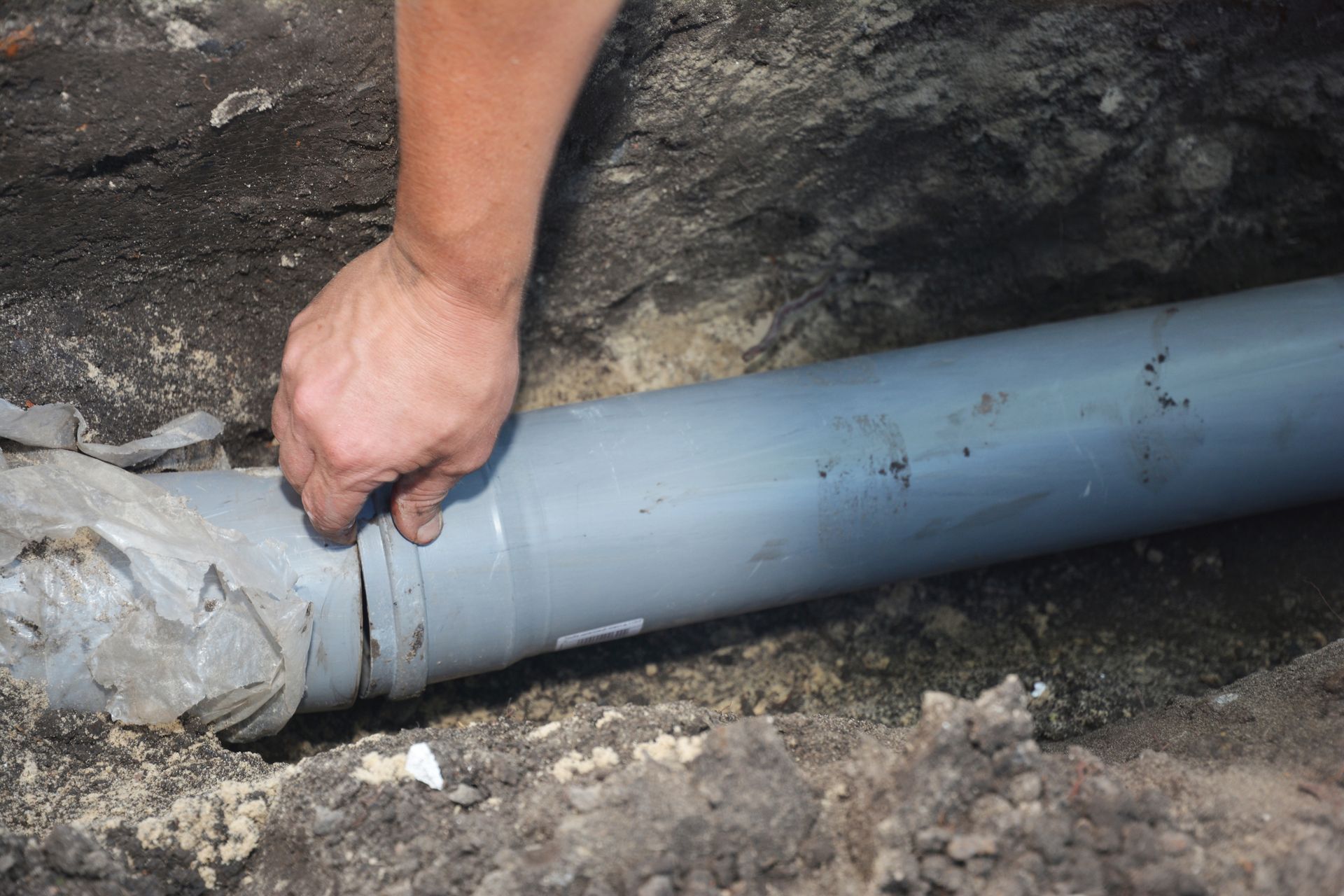 Hand tightening a gray pipe connector in a dirt trench beside a rocky wall