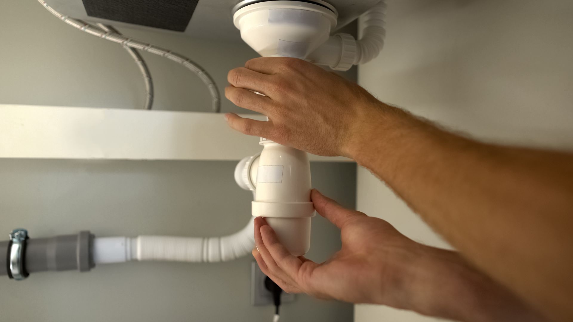 Hands assembling a white sink drain pipe under a countertop