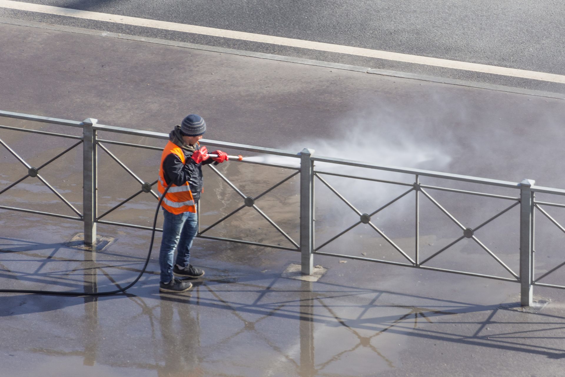 Worker in orange safety vest pressure-washing a wet sidewalk beside a metal railing.