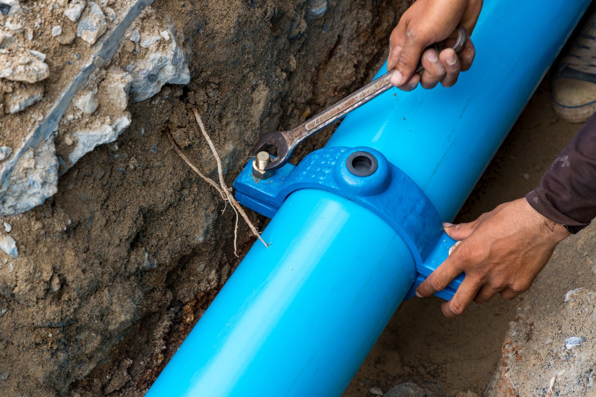 Hands use a wrench to tighten a blue underground pipe joint at a construction site