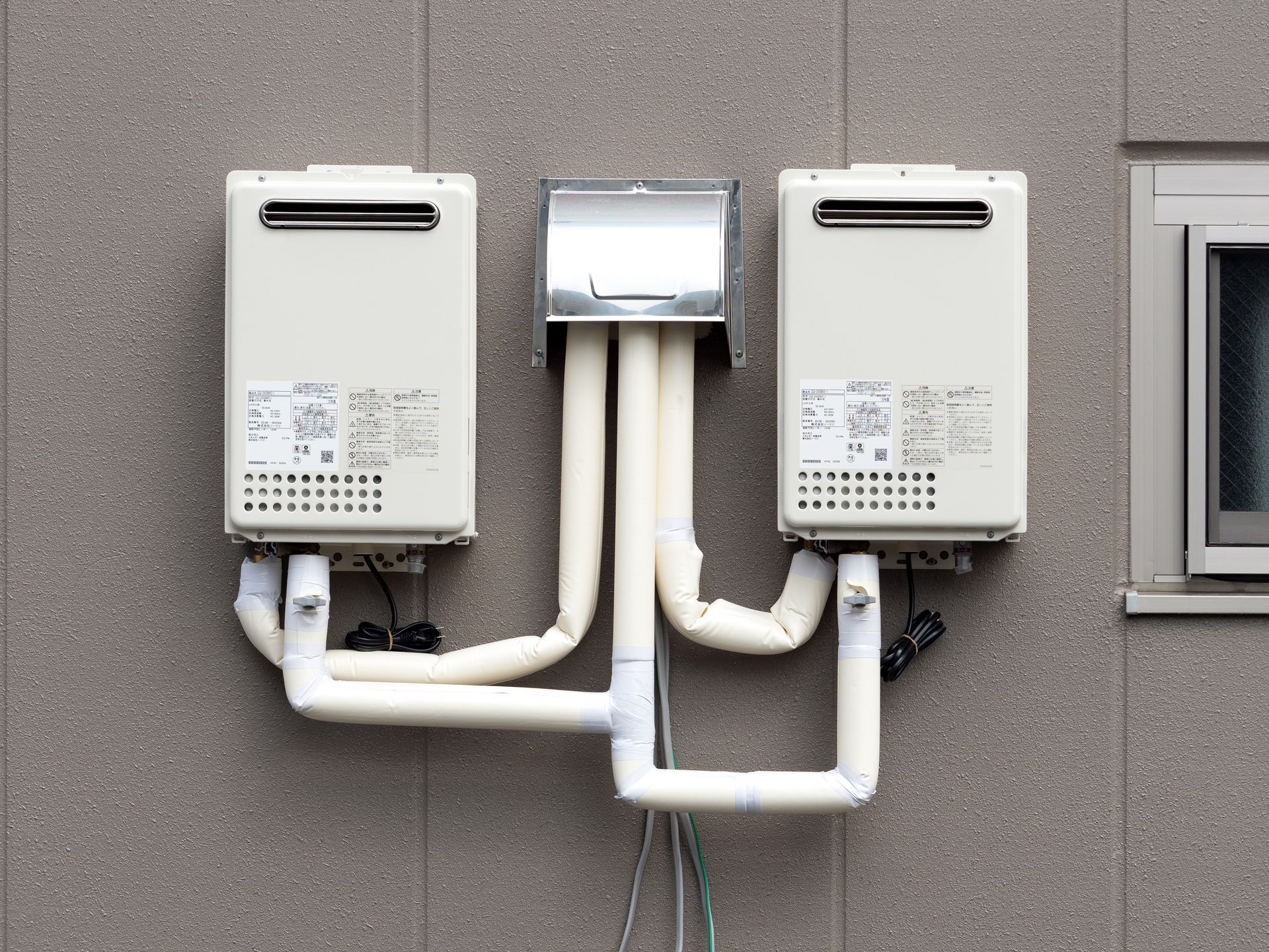 Two beige utility boxes mounted on a wall with white pipes connecting them to a central vent.