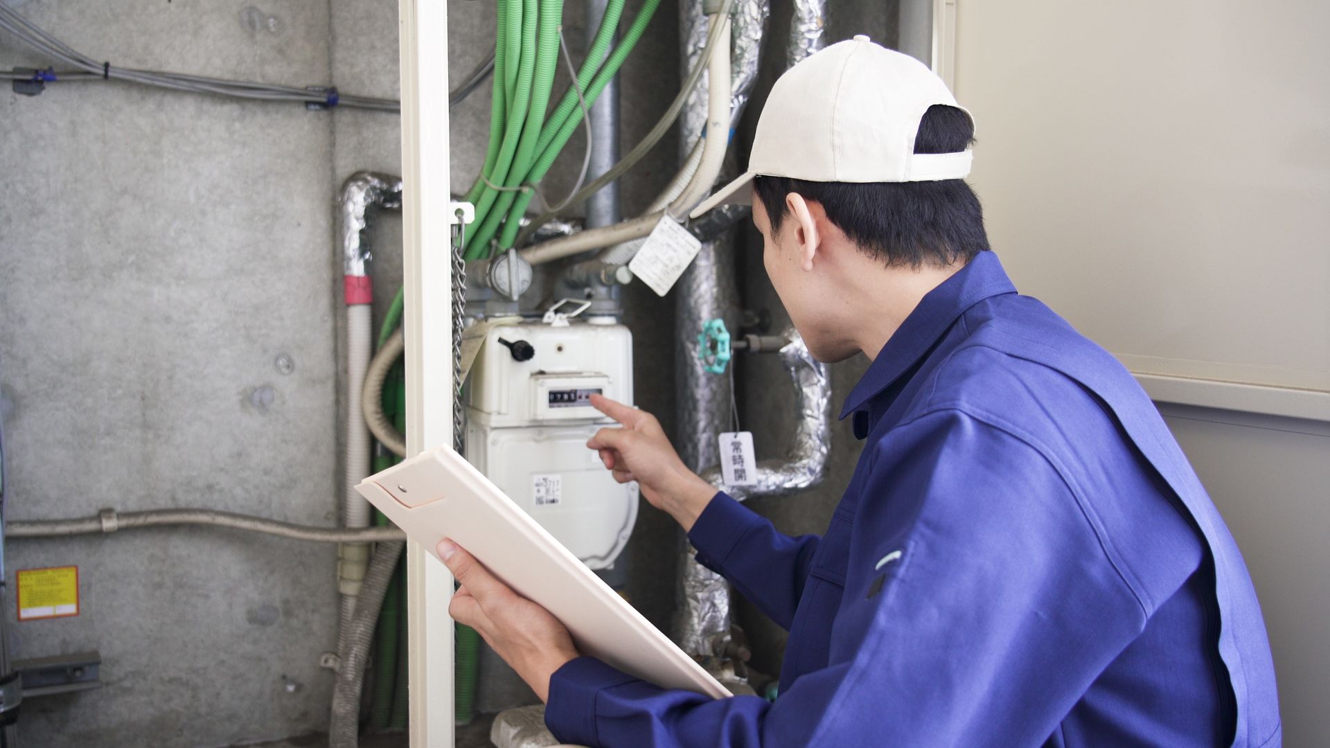 Technician in blue uniform and white cap inspects electrical equipment while holding a clipboard.