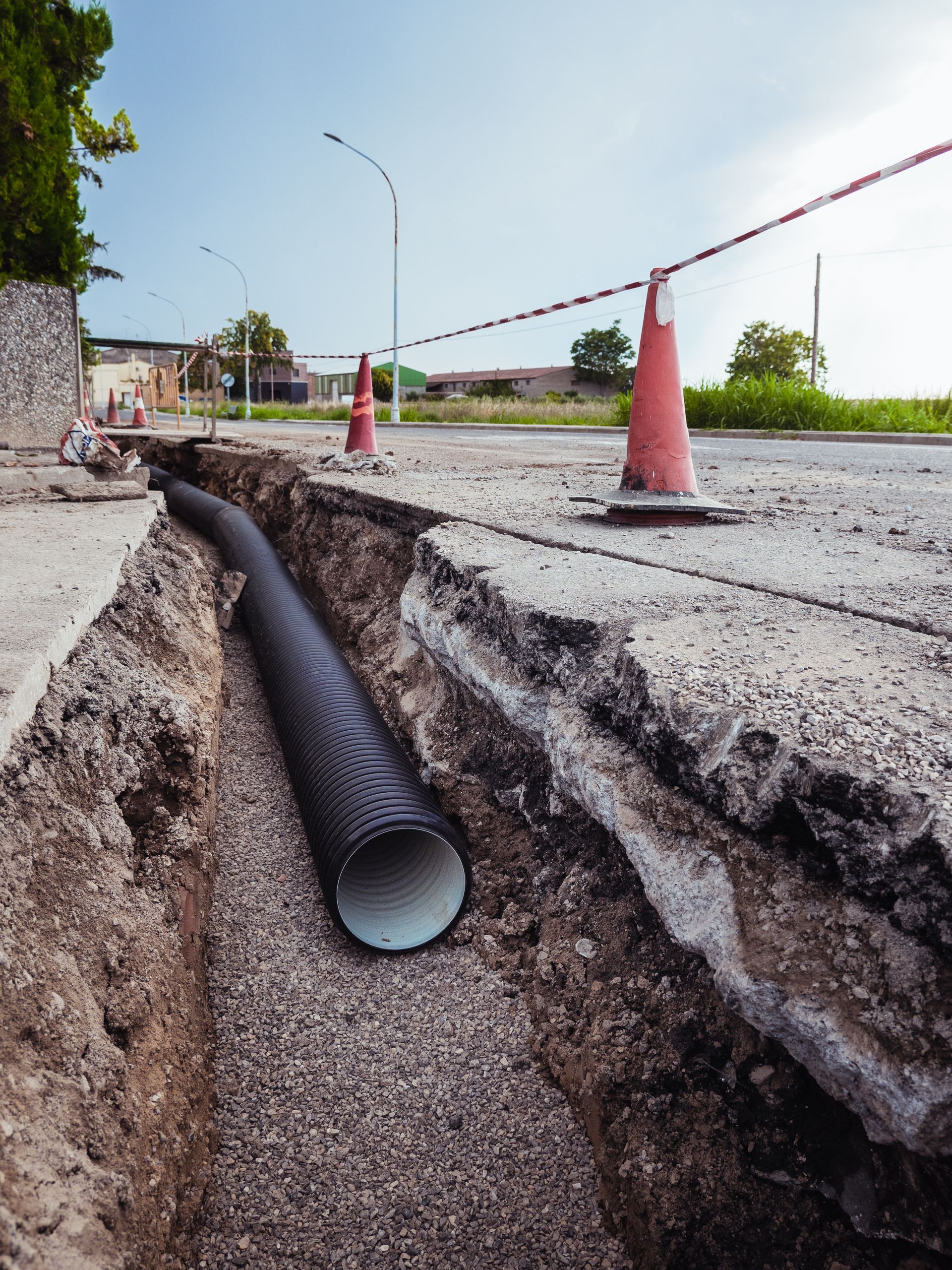 Roadwork trench with a large black pipe, orange cones, and caution tape along a street.