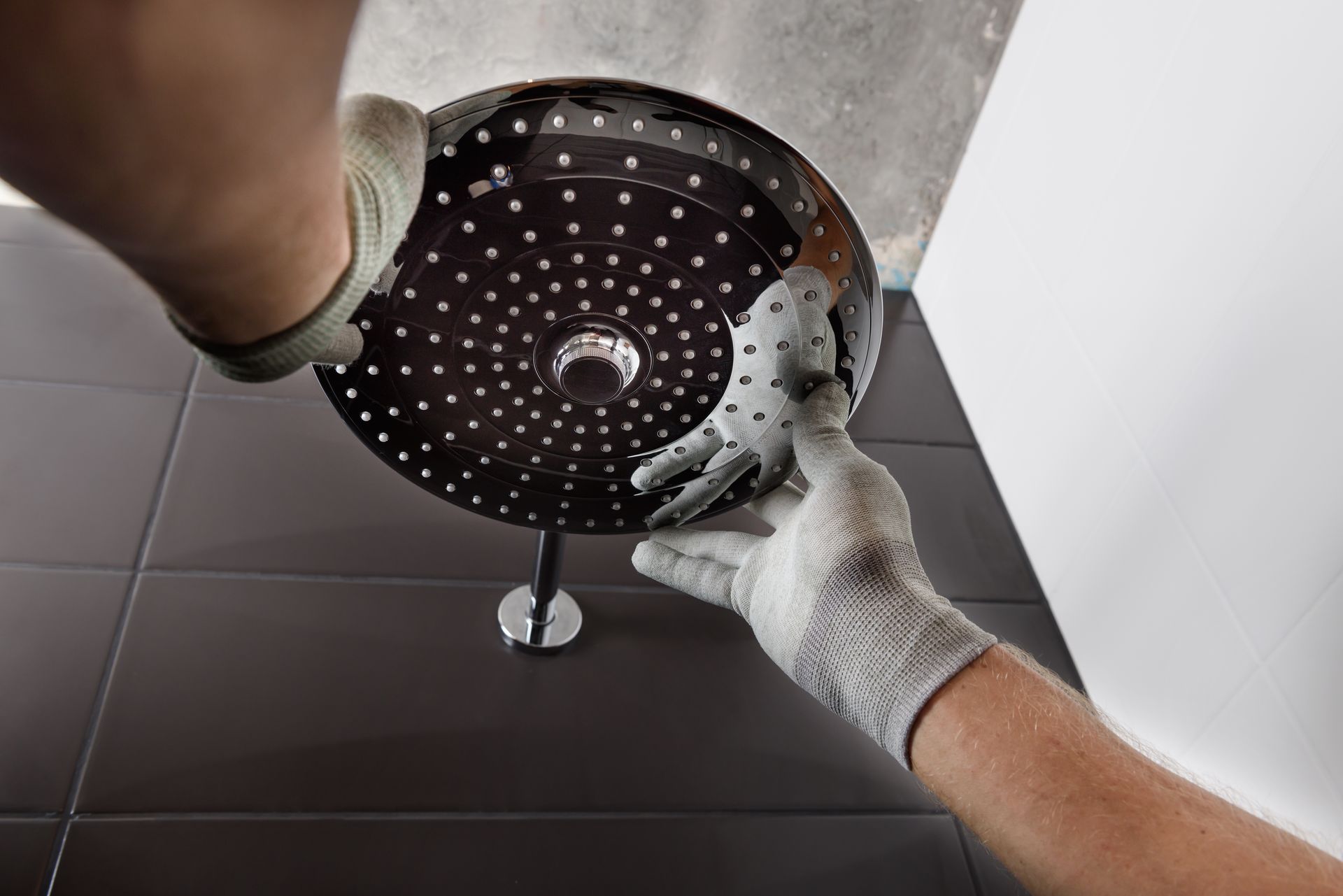 Gloved hands installing a round black shower head against a tiled wall