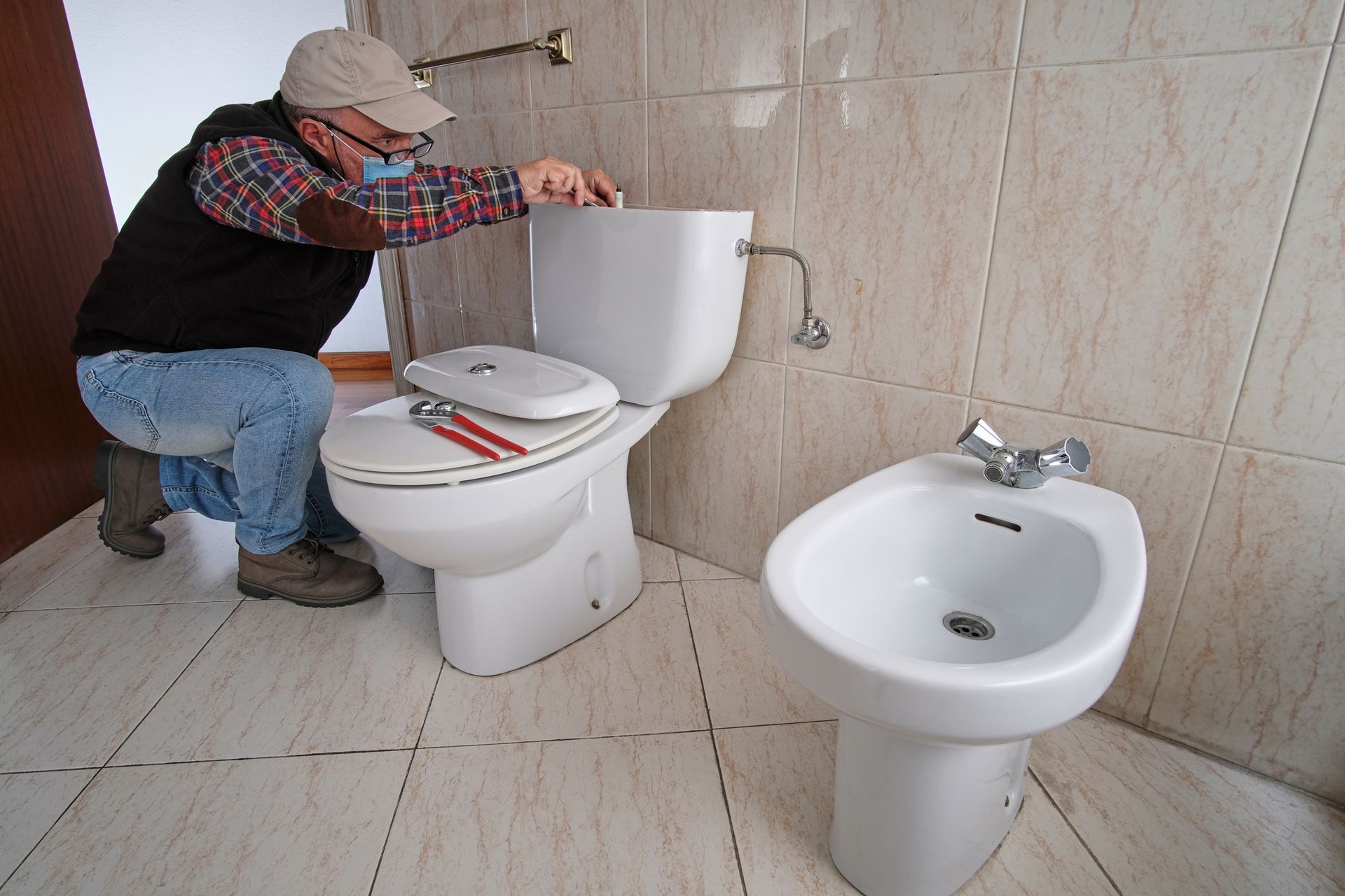 Person kneeling beside a toilet, adjusting plumbing in a tiled bathroom with a sink nearby