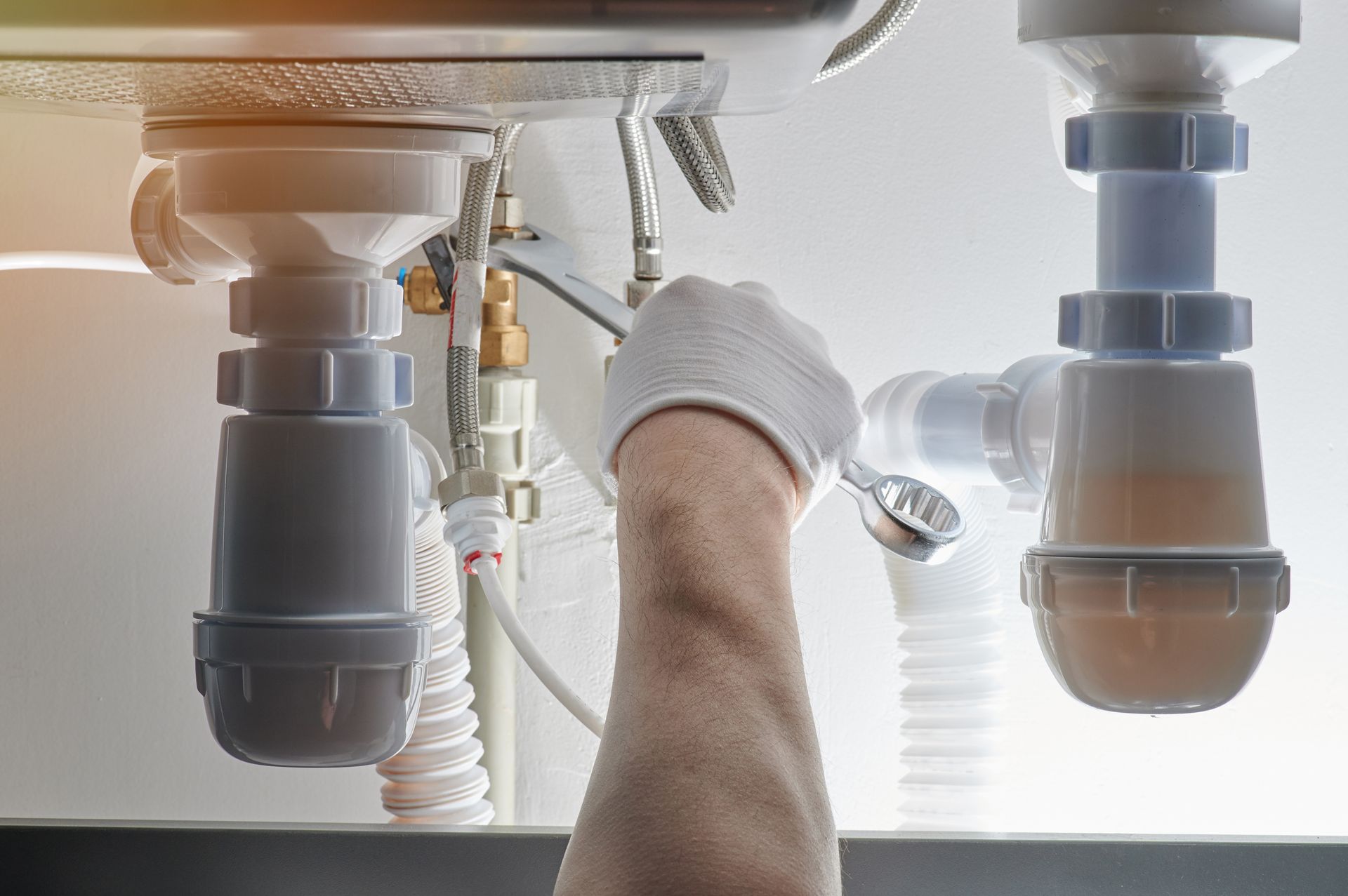 Plumber tightening a sink pipe under a countertop, with gray plumbing and a hand tool visible