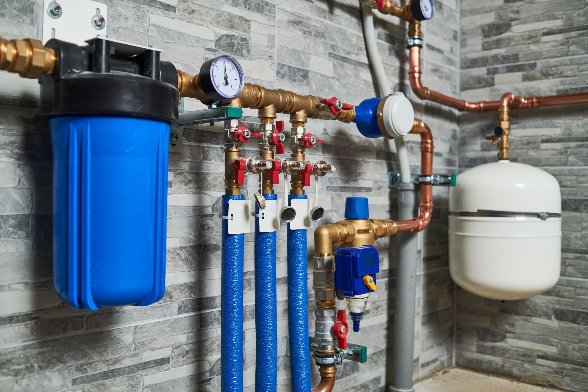 Basement plumbing setup with blue filters, gauges, pipes, and a white expansion tank on a brick wall