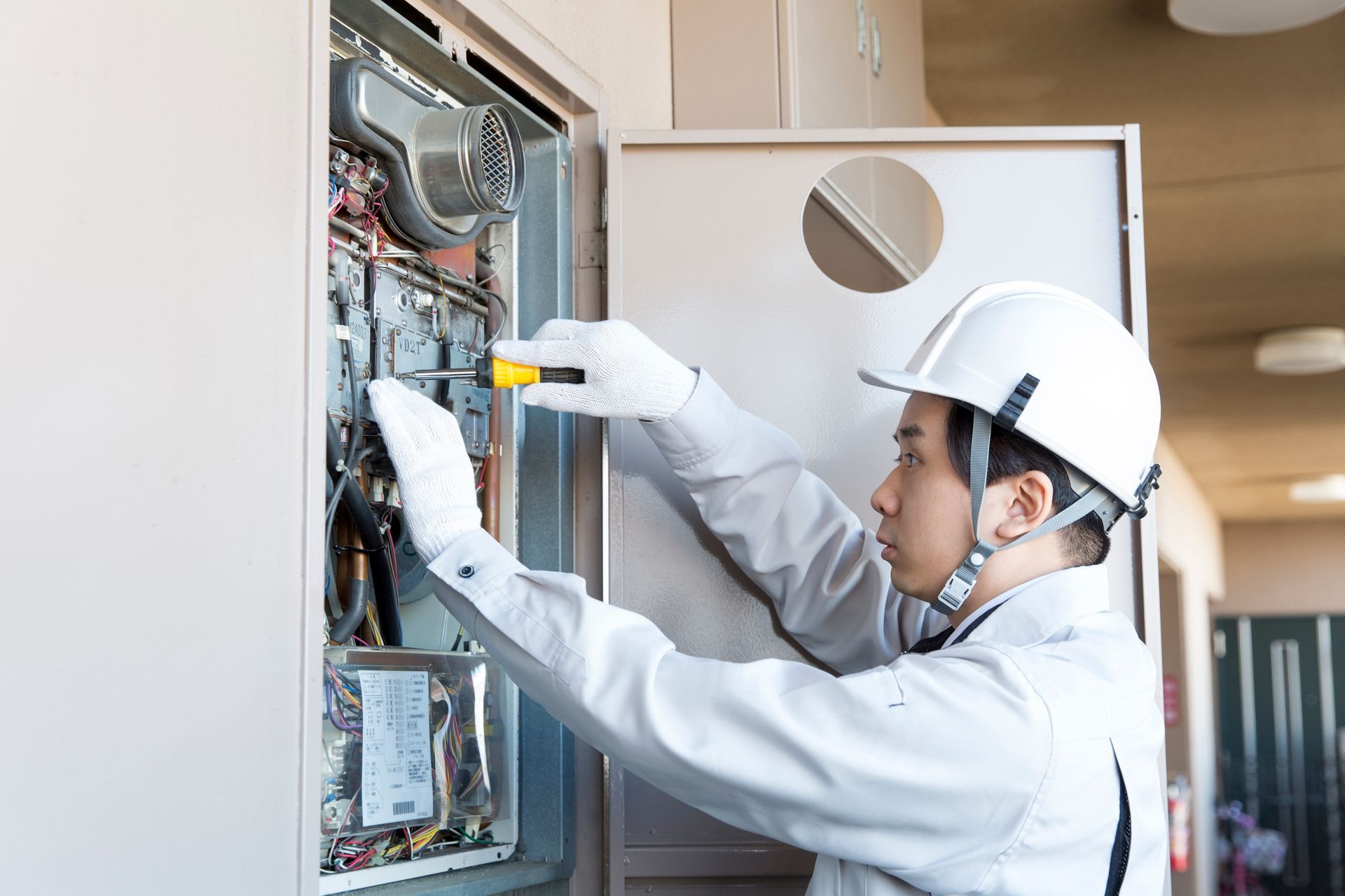 Technician inspecting wiring inside an open electrical panel with a tool, wearing a hard hat and gloves.