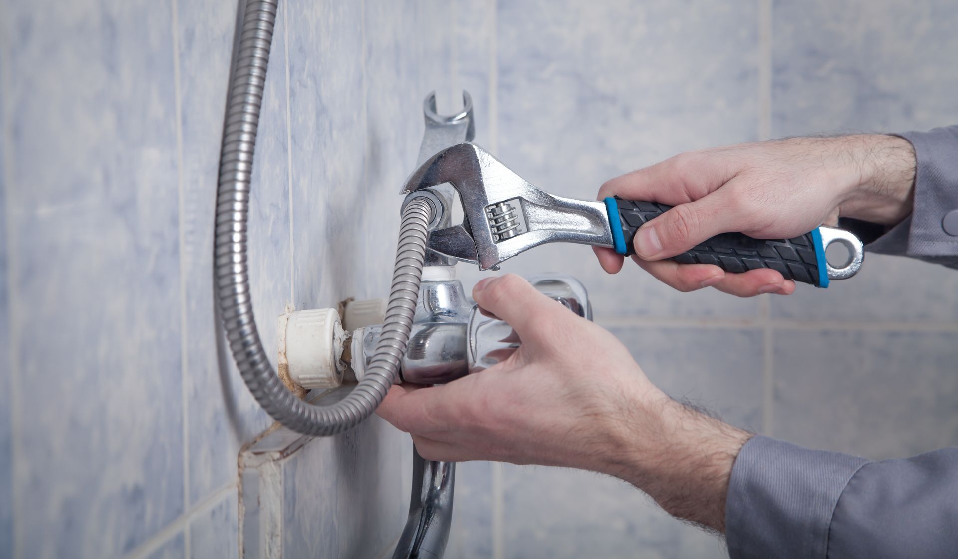 Hands adjusting a chrome shower hose fitting against a tiled wall with a wrench