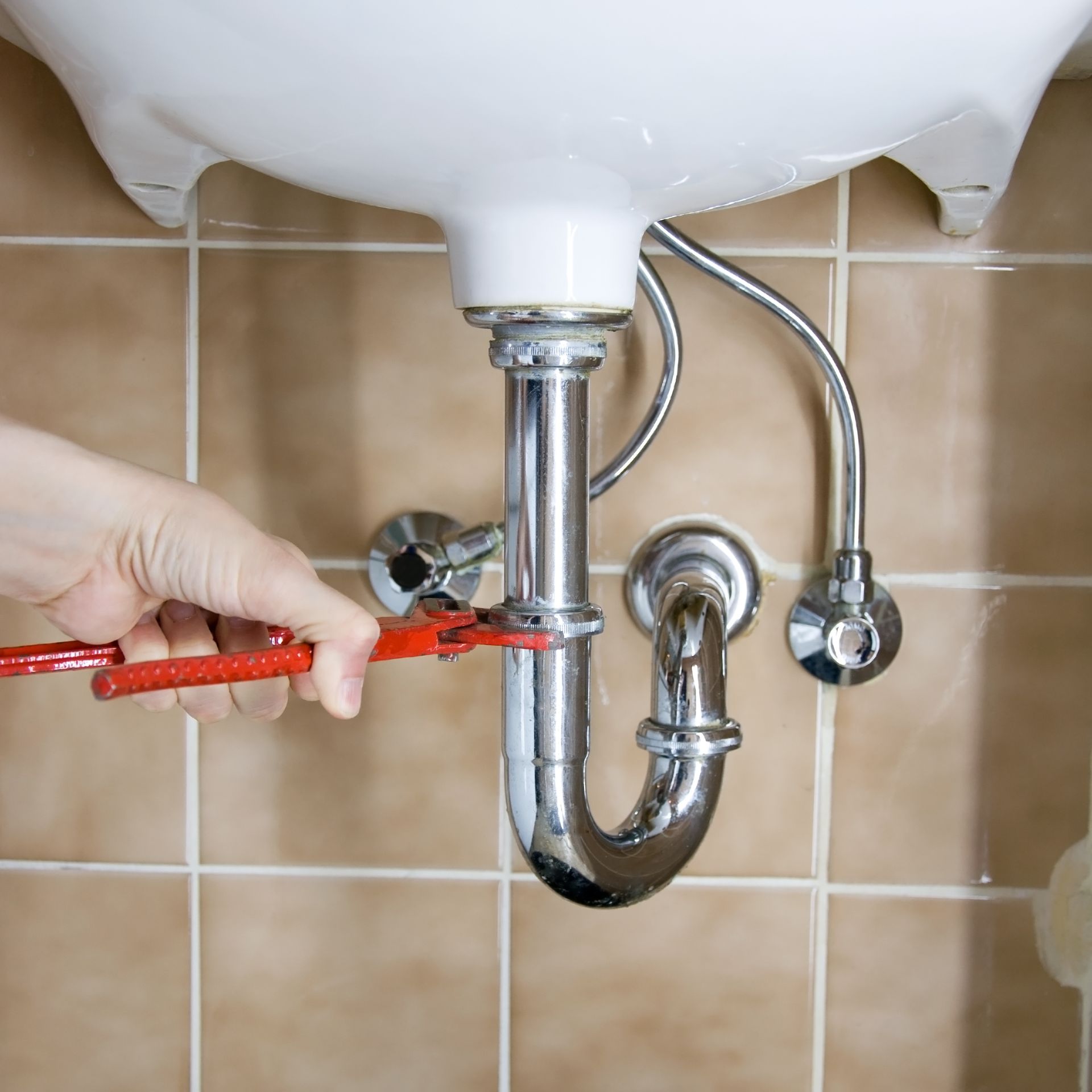 Hand tightening a chrome sink drain pipe with pliers under a white bathroom sink