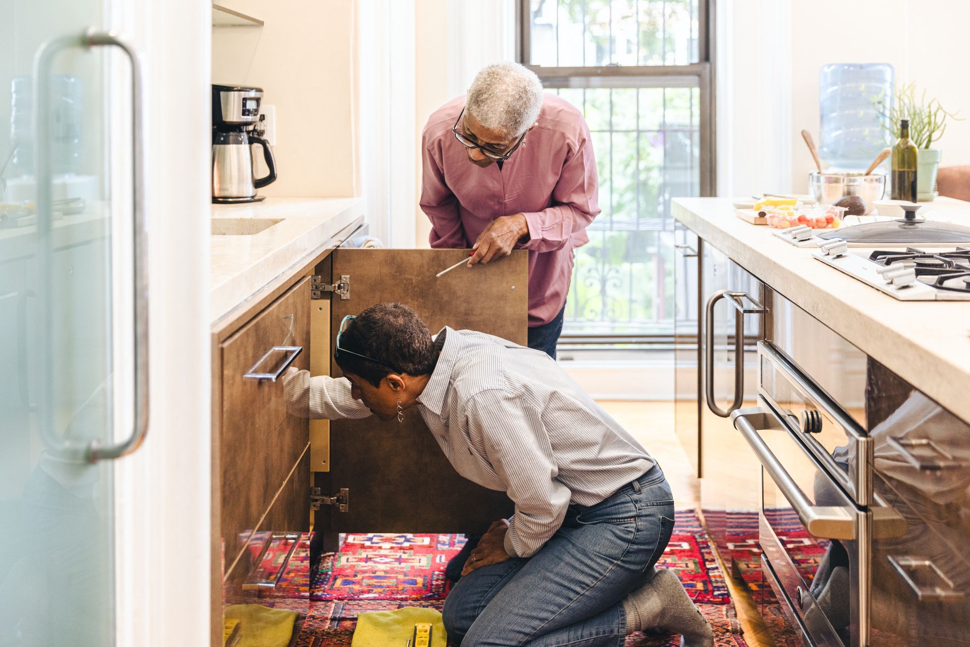 Two people inspect plumbing under a kitchen sink, one kneeling while the other stands nearby.
