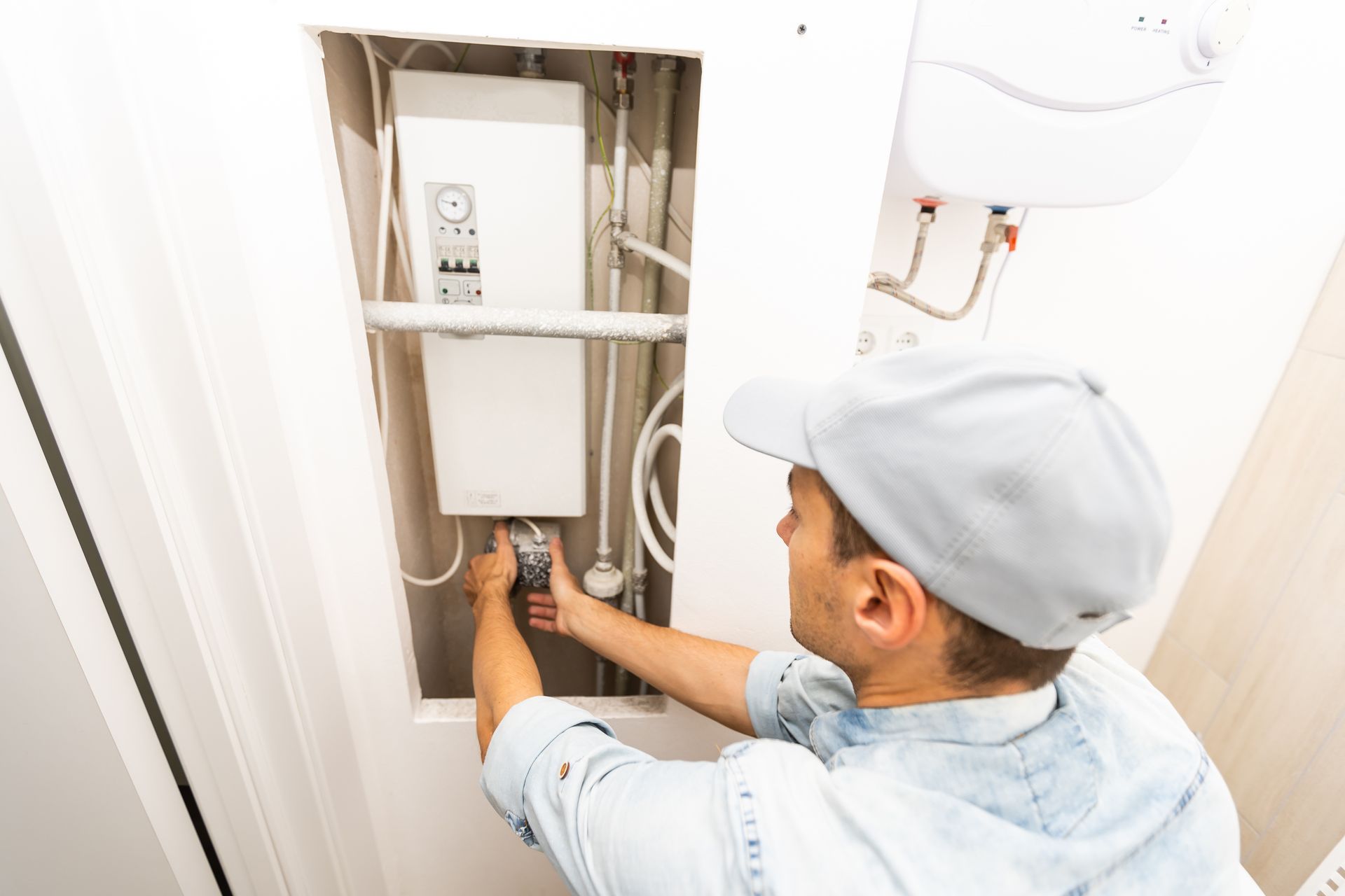 Technician inspecting electrical panel in a wall recess, wearing a cap and working with tools