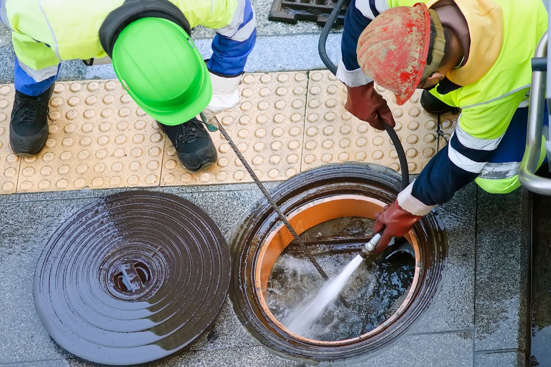 Workers in neon safety gear flushing a manhole with a hose on a street sidewalk