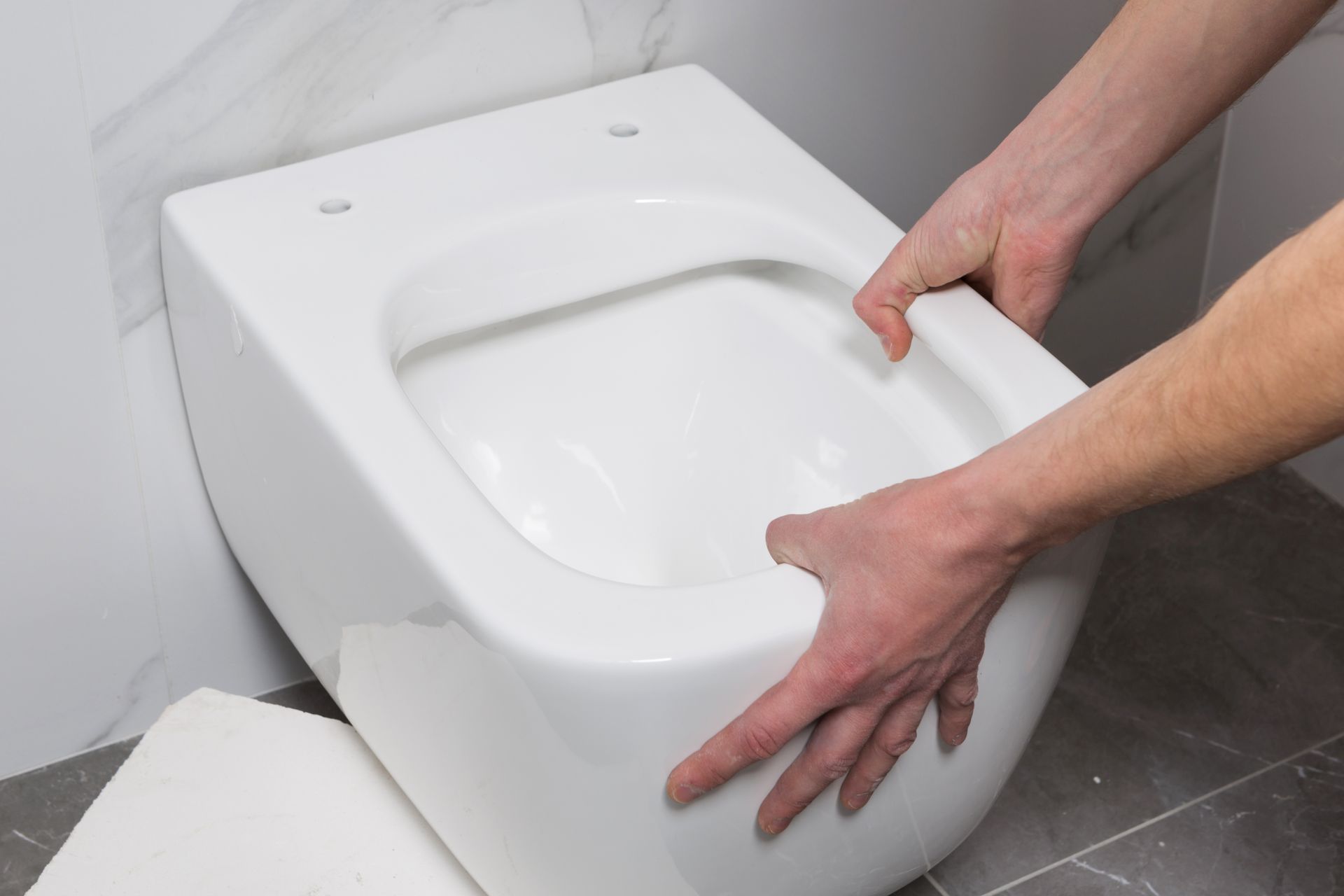Hands installing a white wall-mounted toilet in a bathroom
