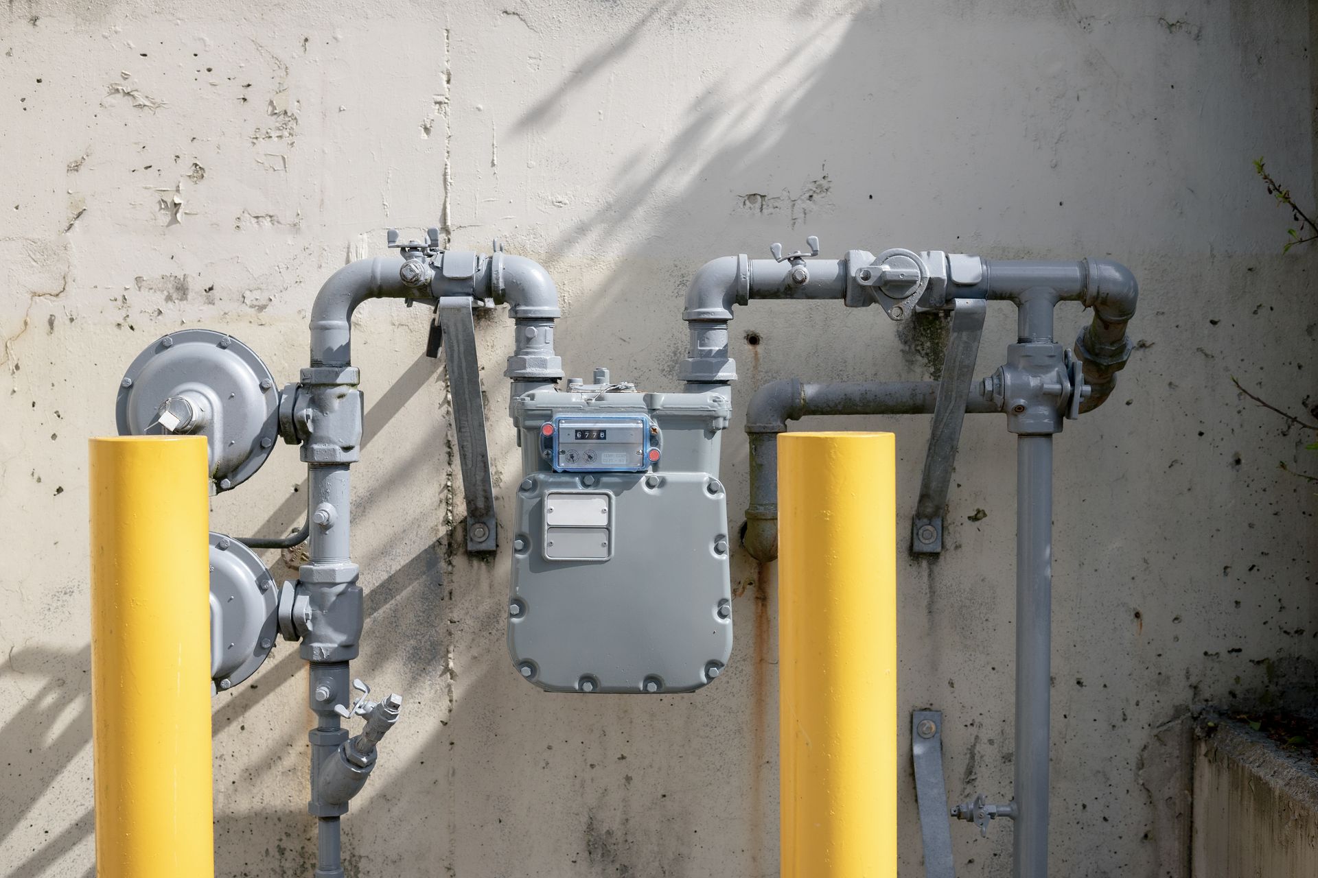Gas meter and pipes mounted on a concrete wall beside yellow safety bollards.